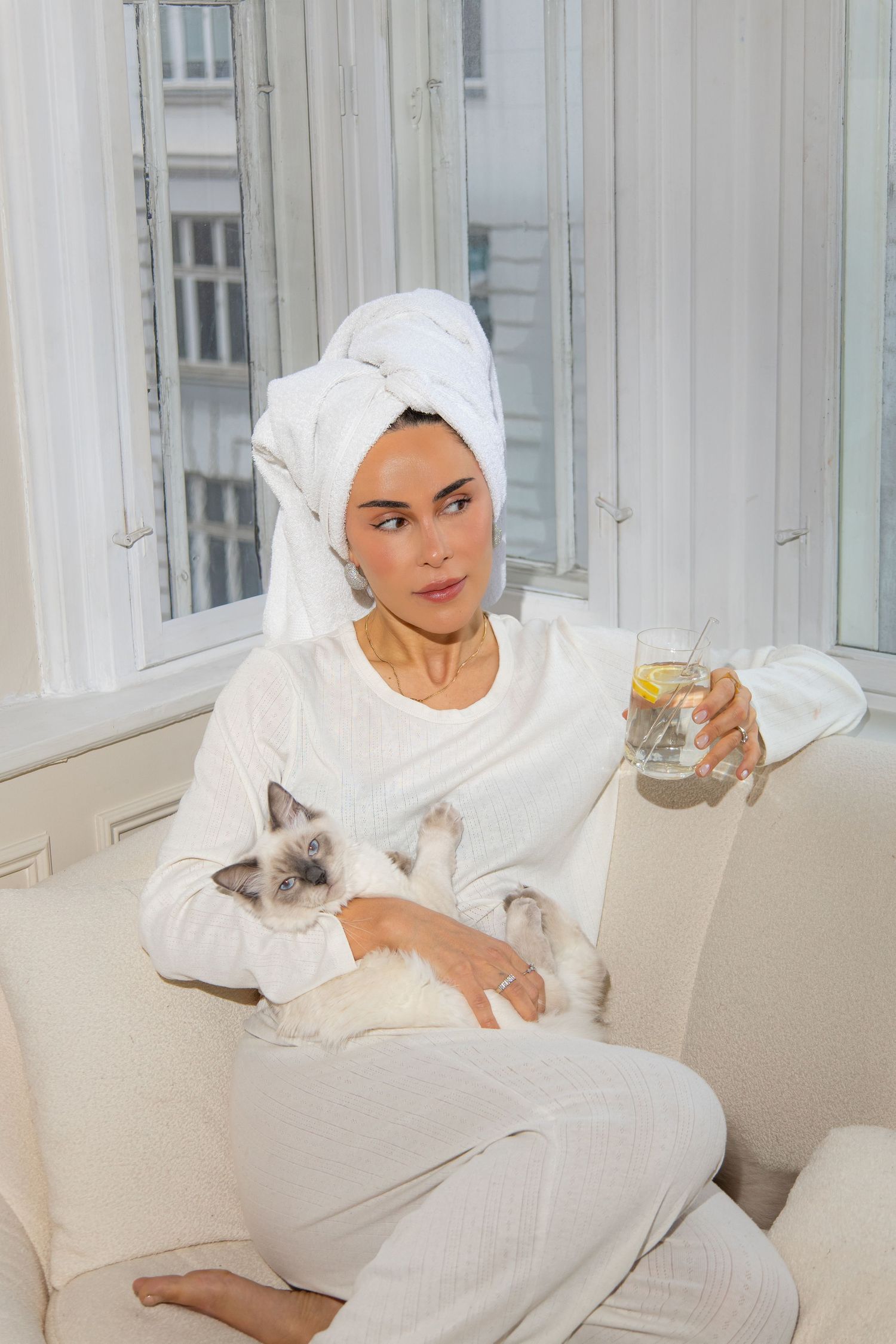 Person in white loungewear relaxes on couch with Siamese cat while enjoying a warm beverage.
