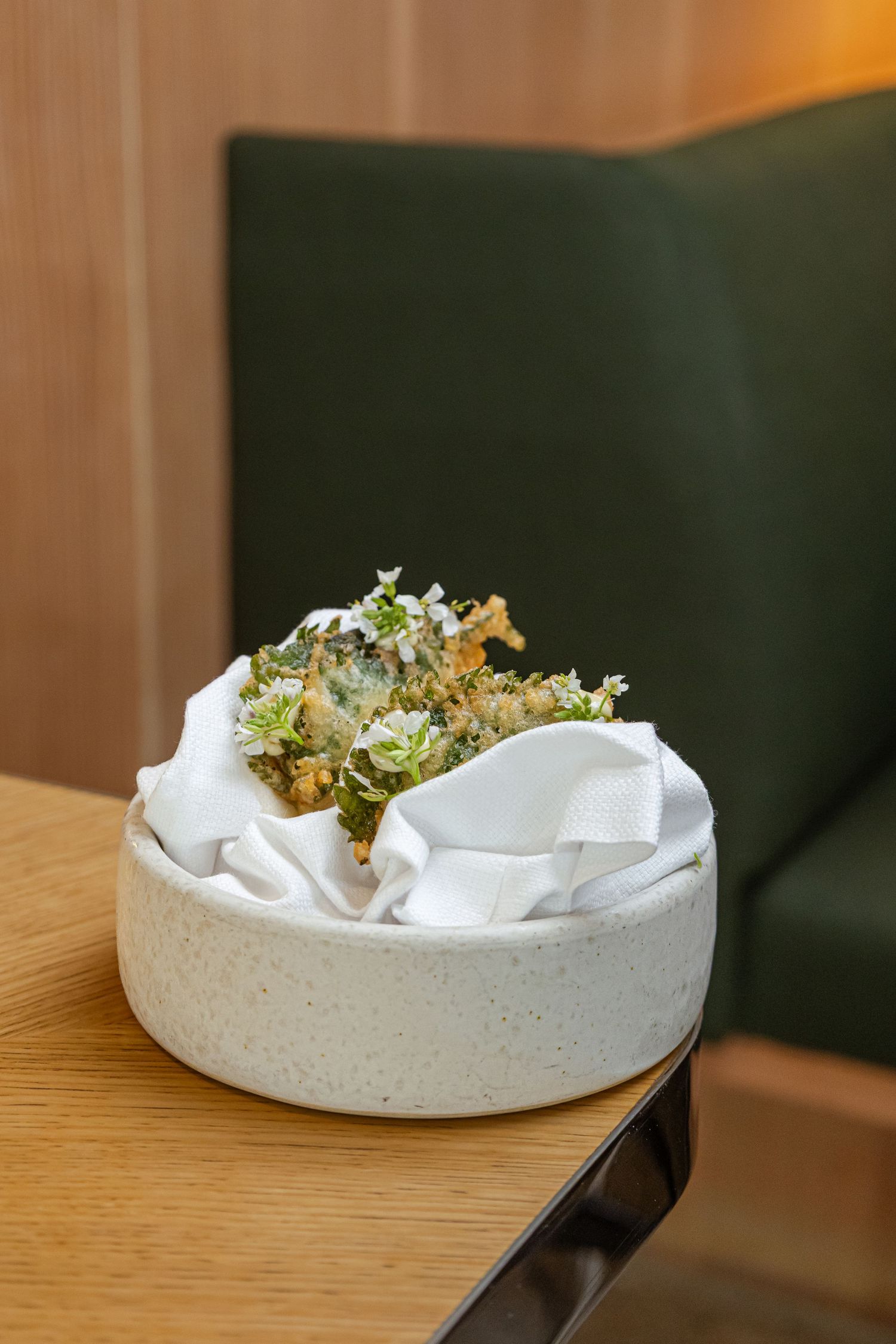 Crispy tempura vegetables served in a white stone bowl with linen napkin on a wooden table in an elegant restaurant.