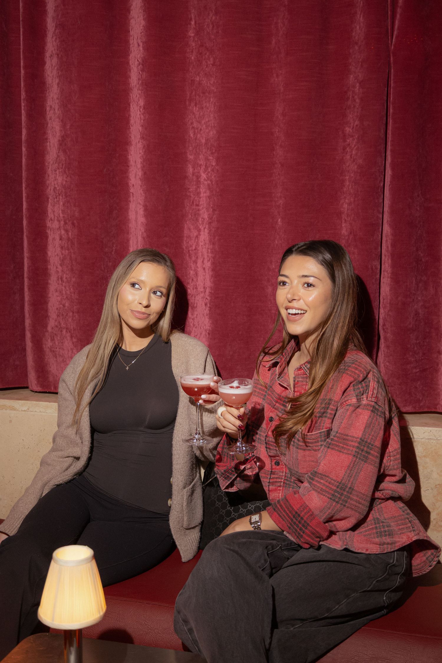 Two women smiling and holding pink cocktails while sitting on a red booth seat in a dimly lit bar.