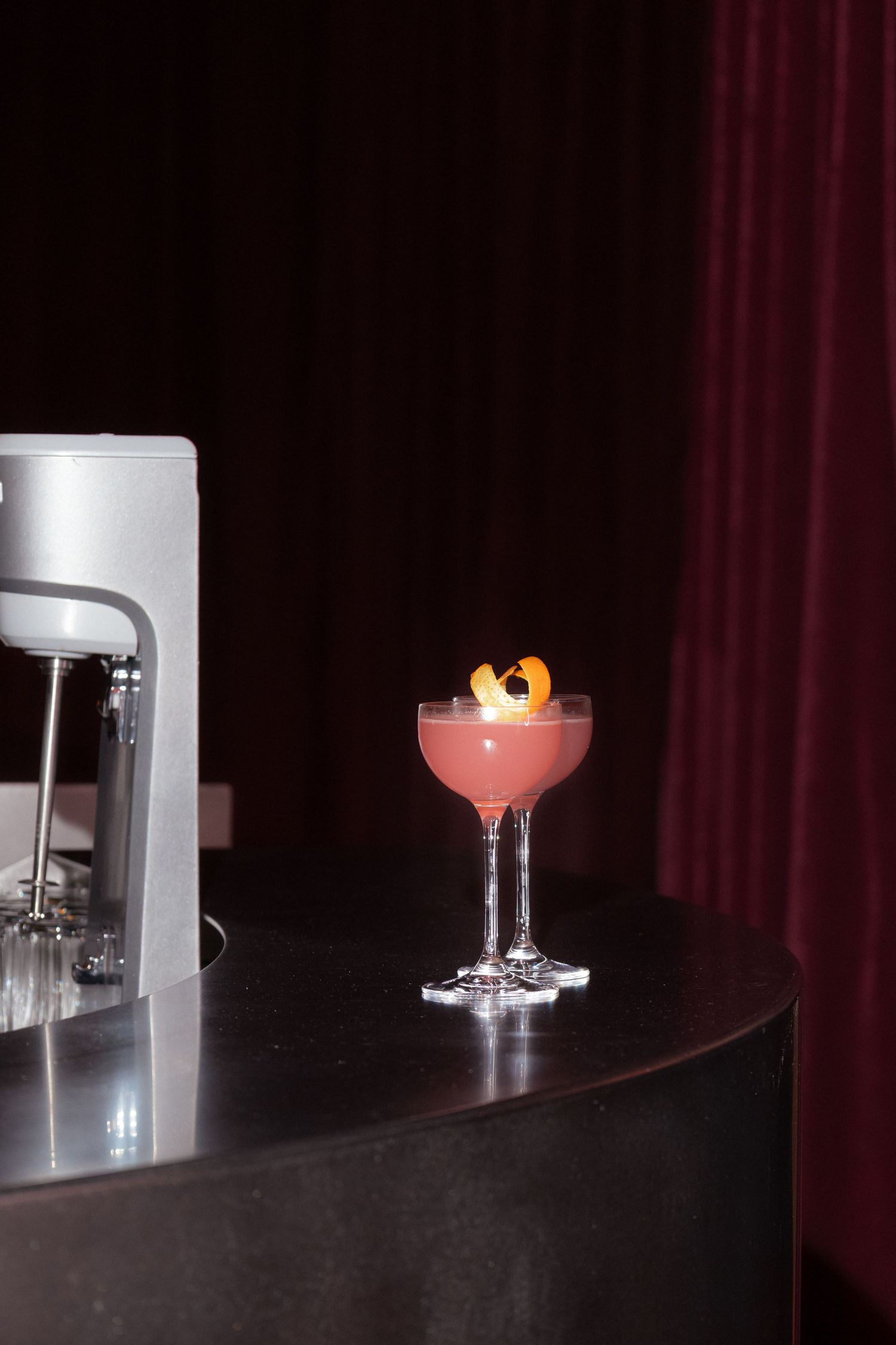 Pink cocktail in coupe glass with citrus garnish on dark bar counter, deep red curtain backdrop.