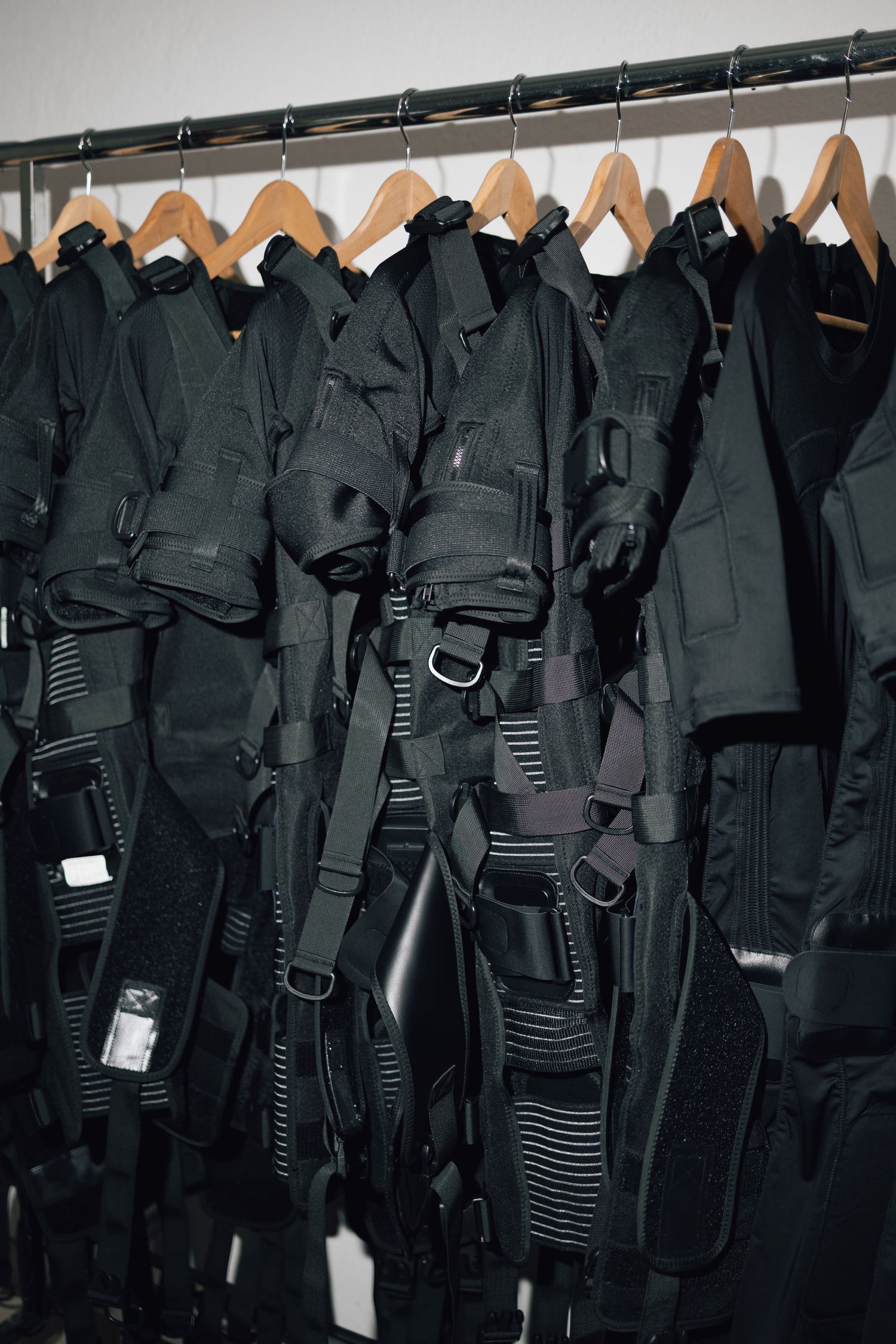 Row of black tactical vests with straps and harnesses hanging on wooden hangers on a clothing rack.