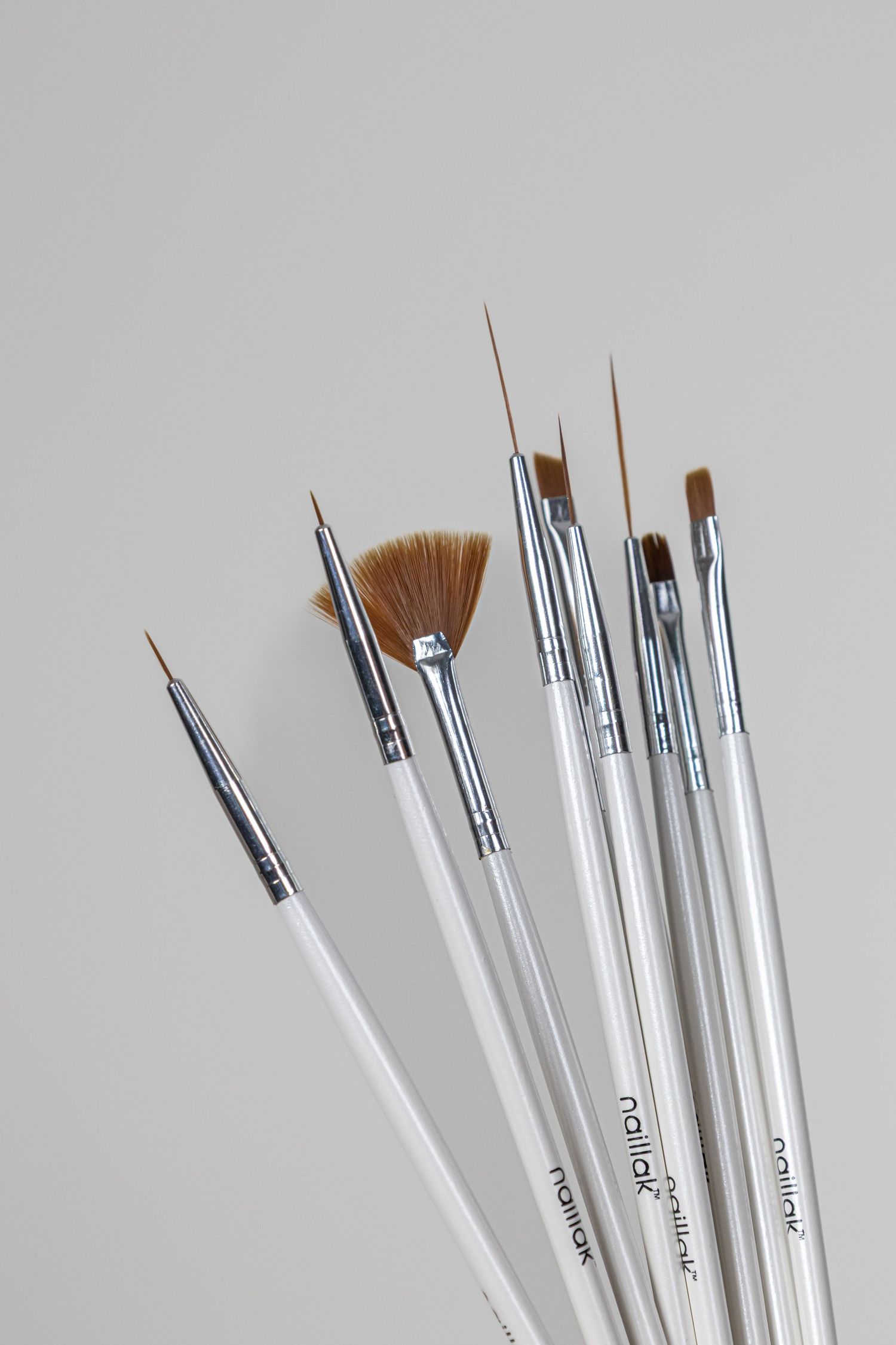 Set of white-handled nail art brushes by Naillak, including fan, liner, and detail brushes on gray background.
