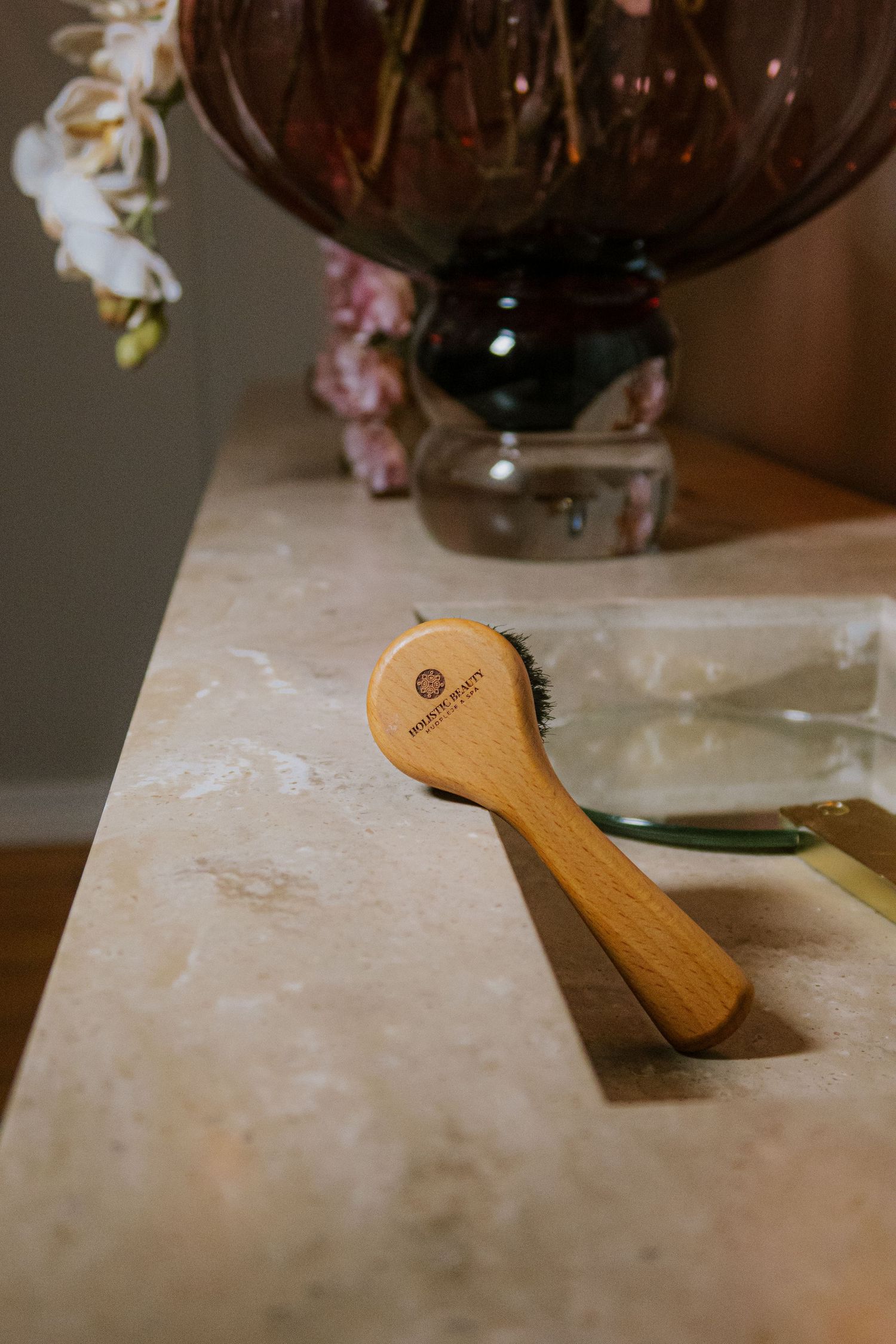 Wooden face brush with bristles resting on marble surface beside glass vase with flowers.