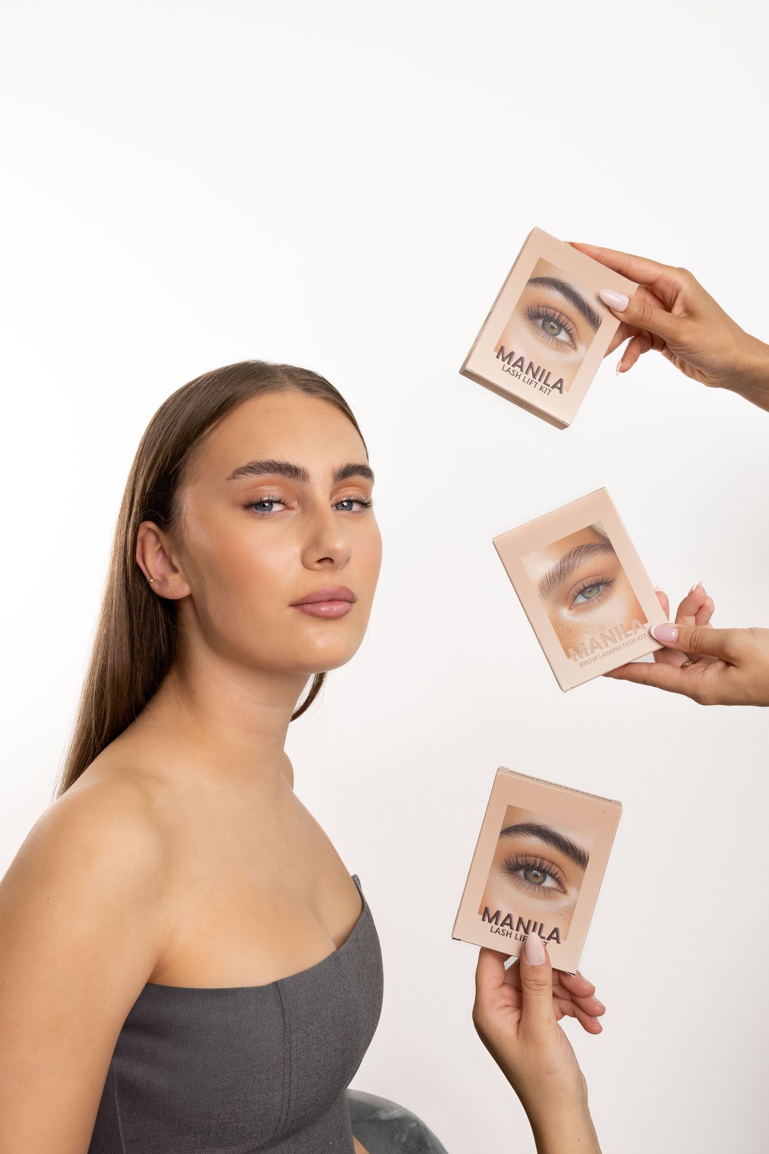 Woman in gray top poses with multiple Manila Lash Lift Kit boxes held around her by hands.