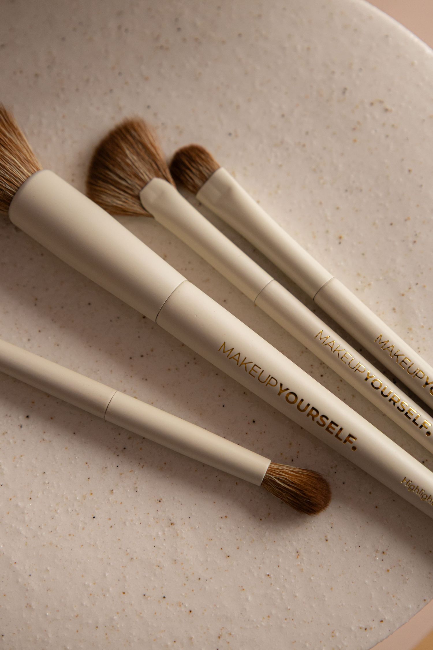 Four MAKEUP YOURSELF cream makeup brushes with brown bristles arranged on a speckled ceramic plate.