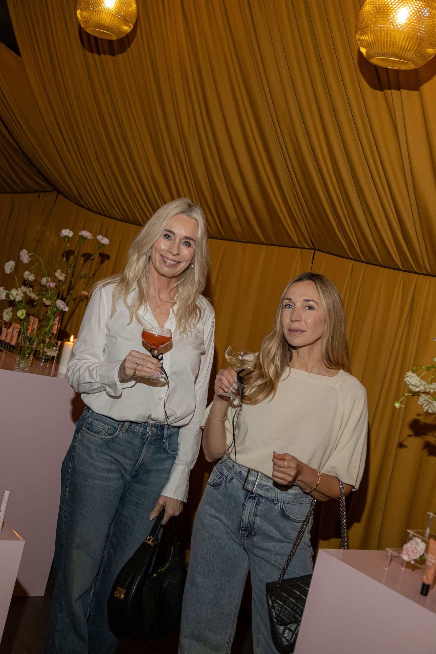 Two blonde women holding wine glasses at an elegant event with gold draped ceiling and floral decor.