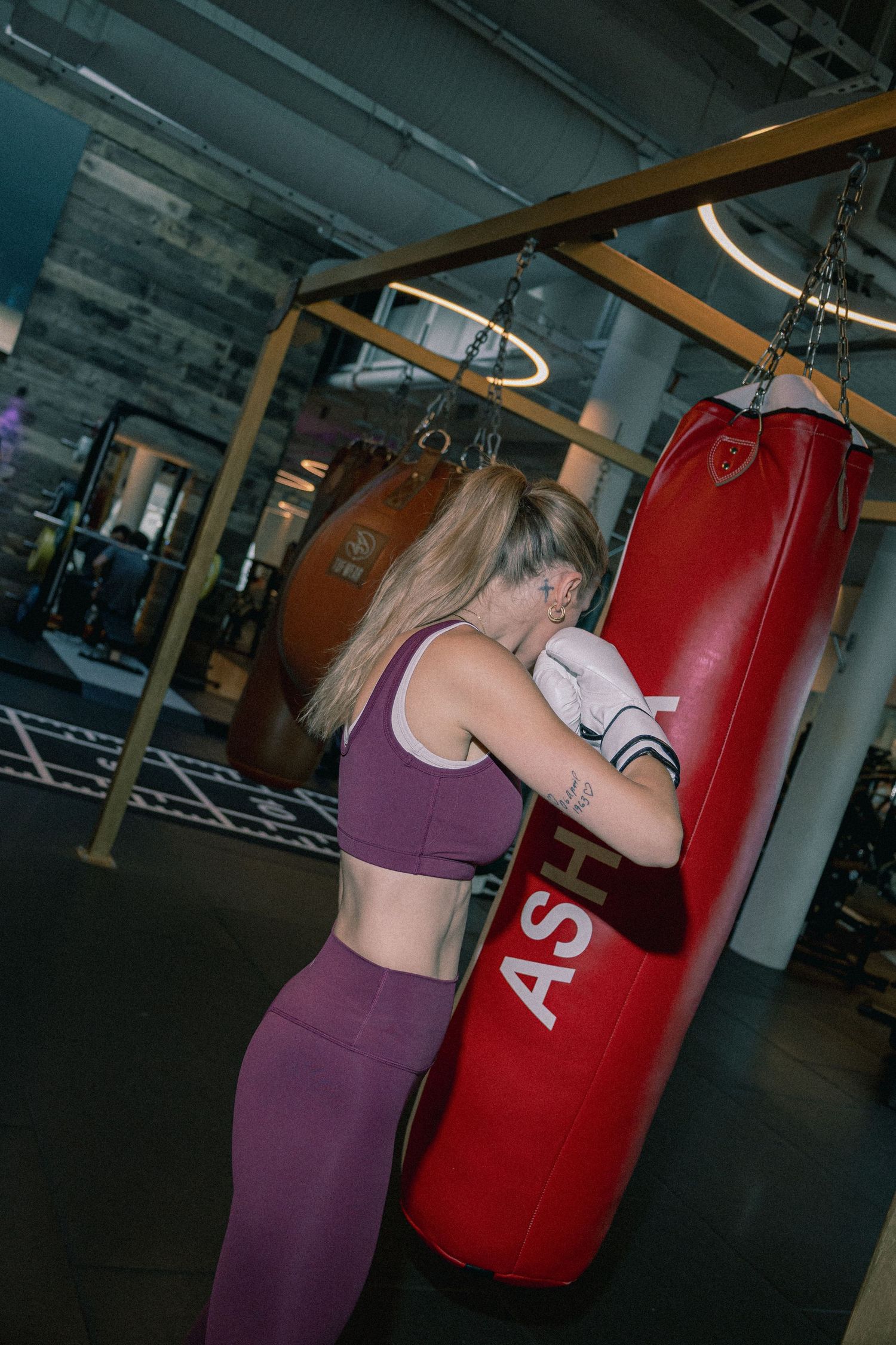 Woman in purple activewear punching a red ASH heavy bag with boxing wraps in a modern gym.