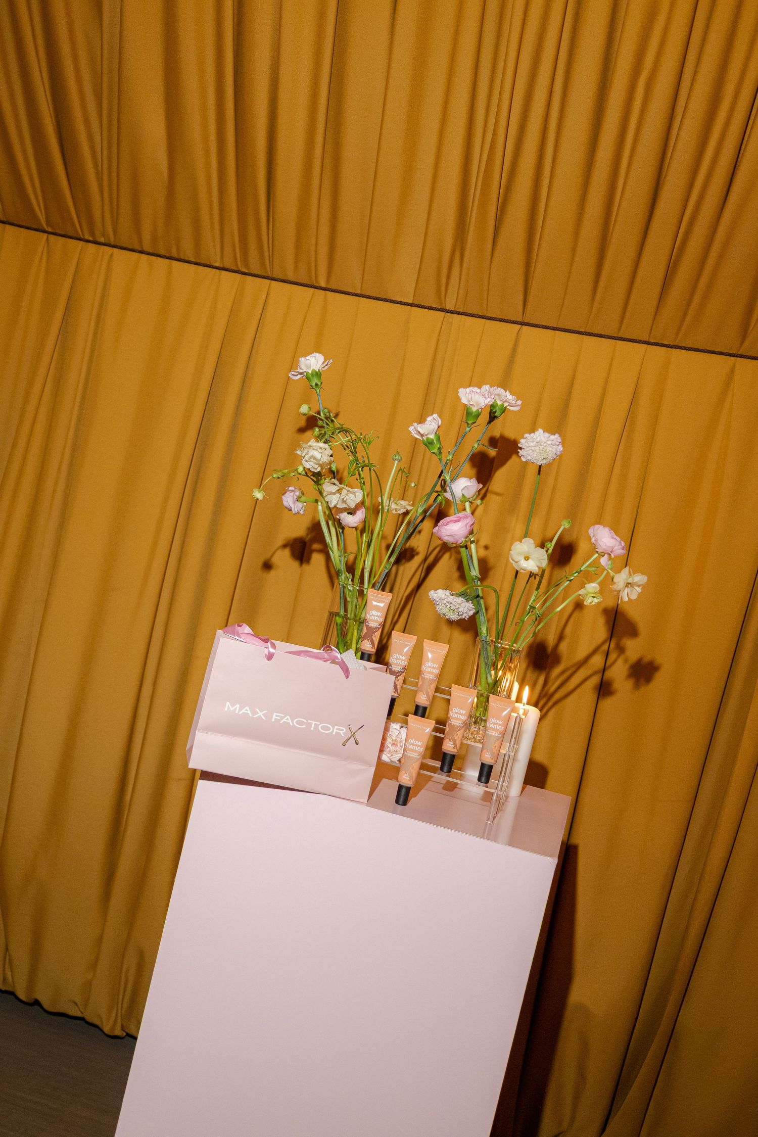 Max Factor beauty products displayed with delicate white and pink flowers in glass vases on a white pedestal against golden curtains.
