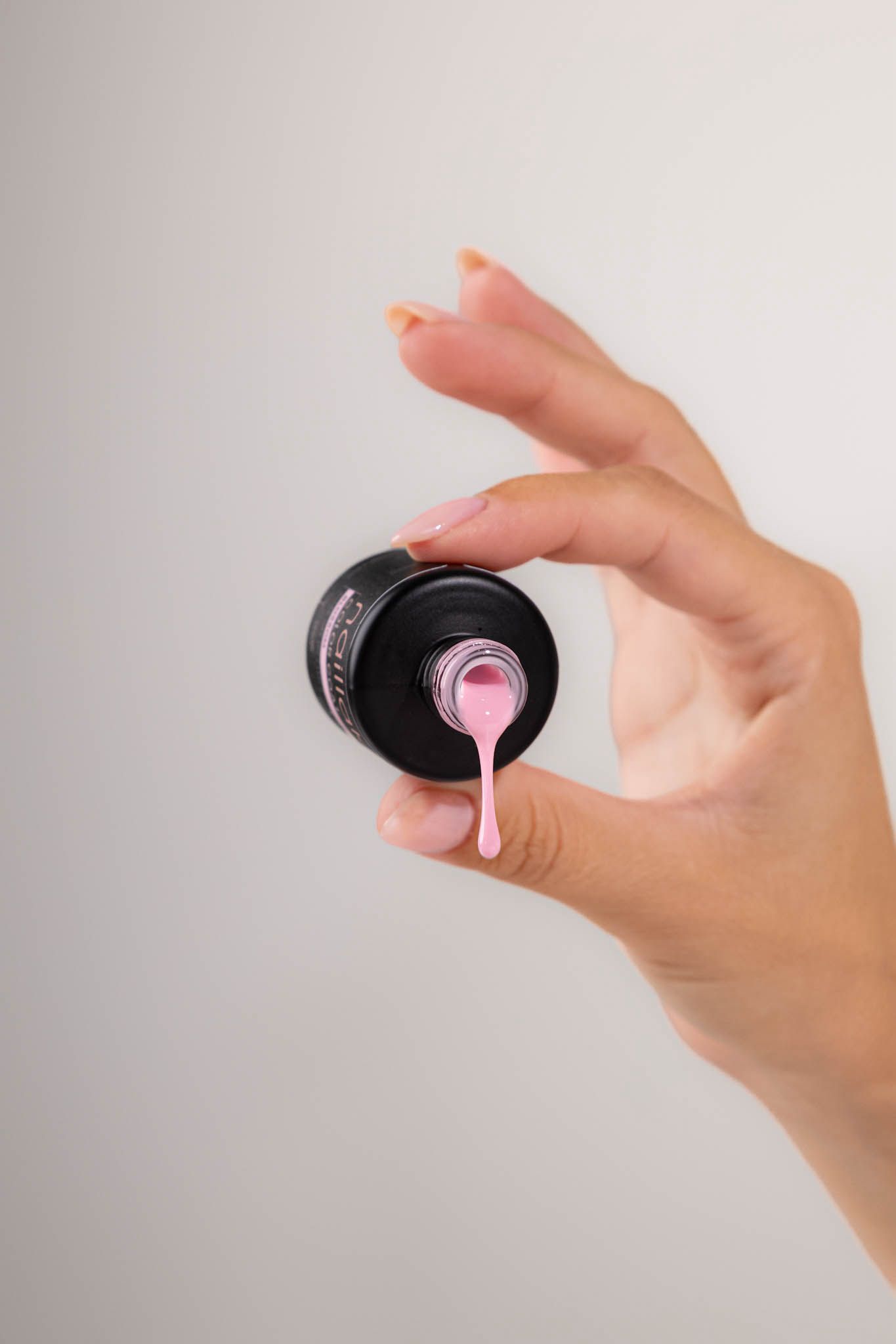A hand holding an inverted black nail polish bottle with pink polish dripping from the brush applicator.