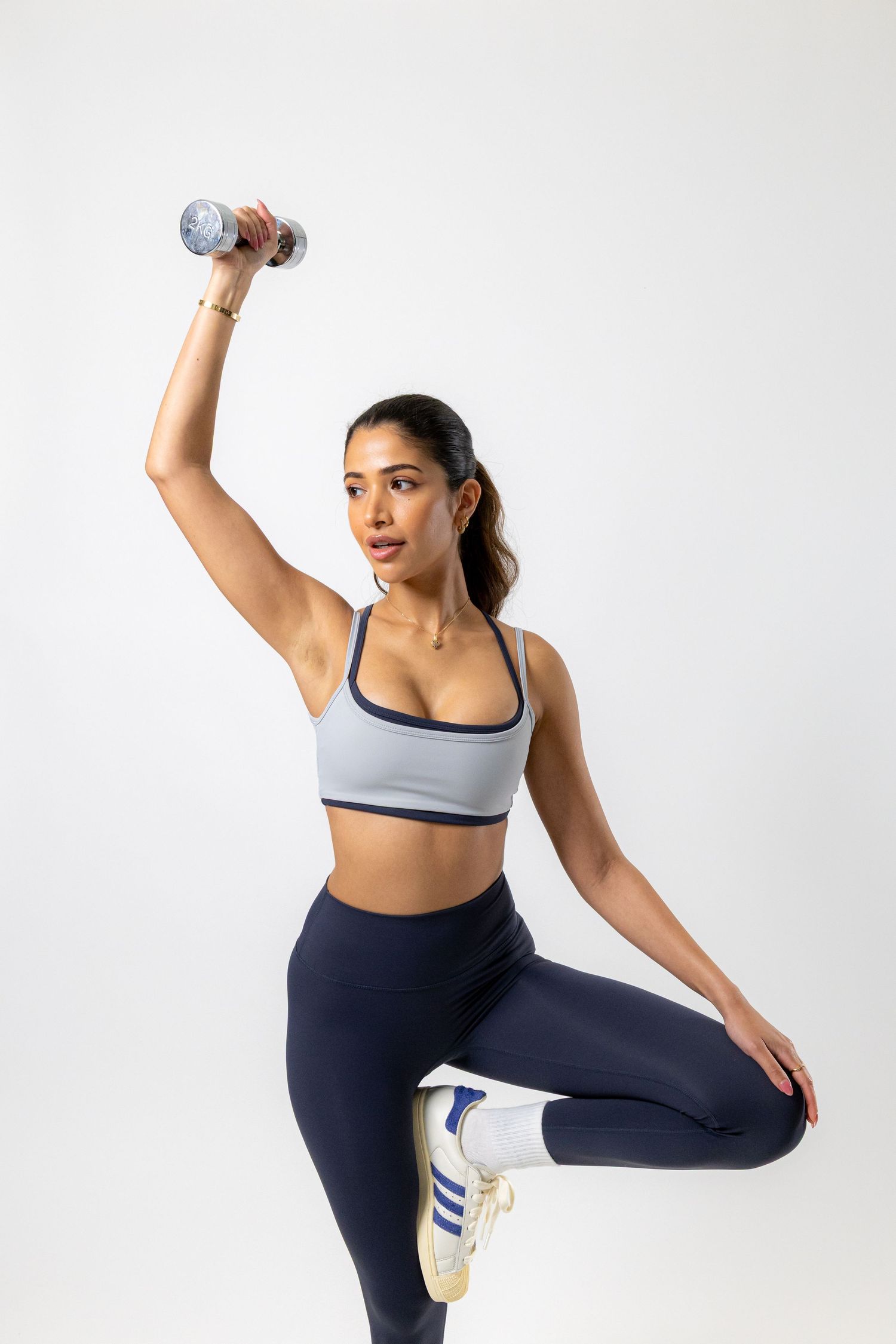 Woman in gray sports bra and navy leggings raises dumbbell overhead while balancing on one leg against white background.