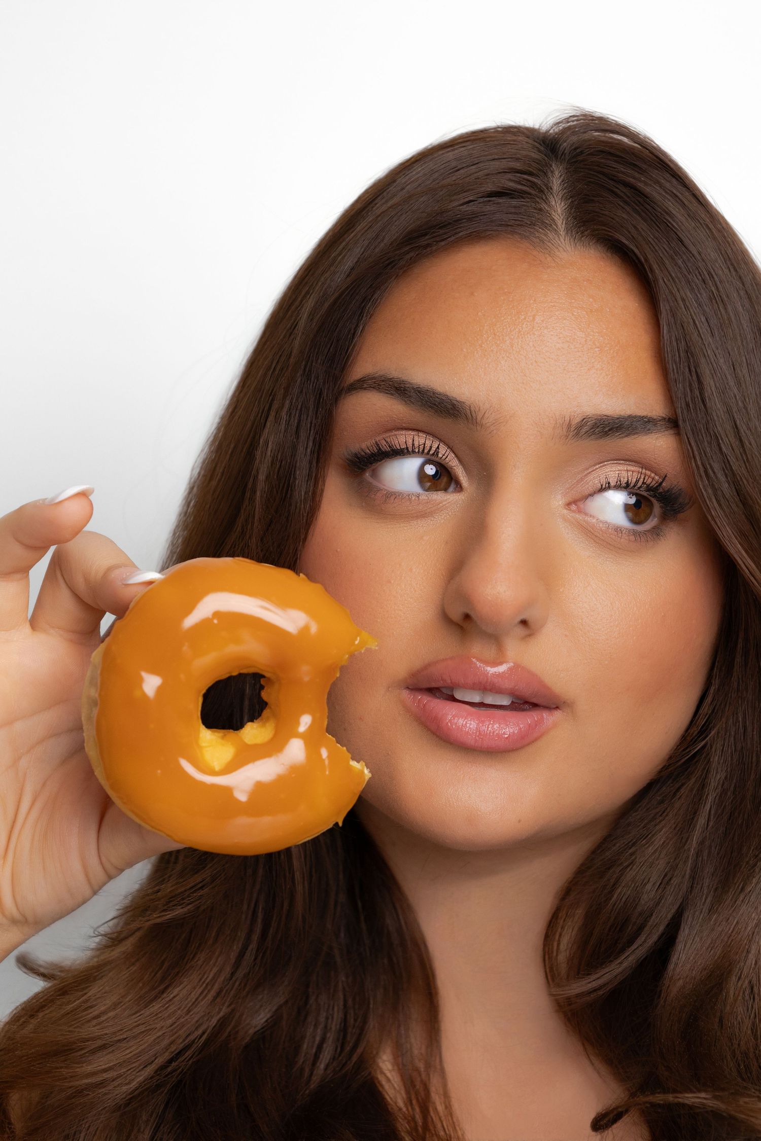 Young woman with brown eyes holding a glazed orange donut near her face against a white background.
