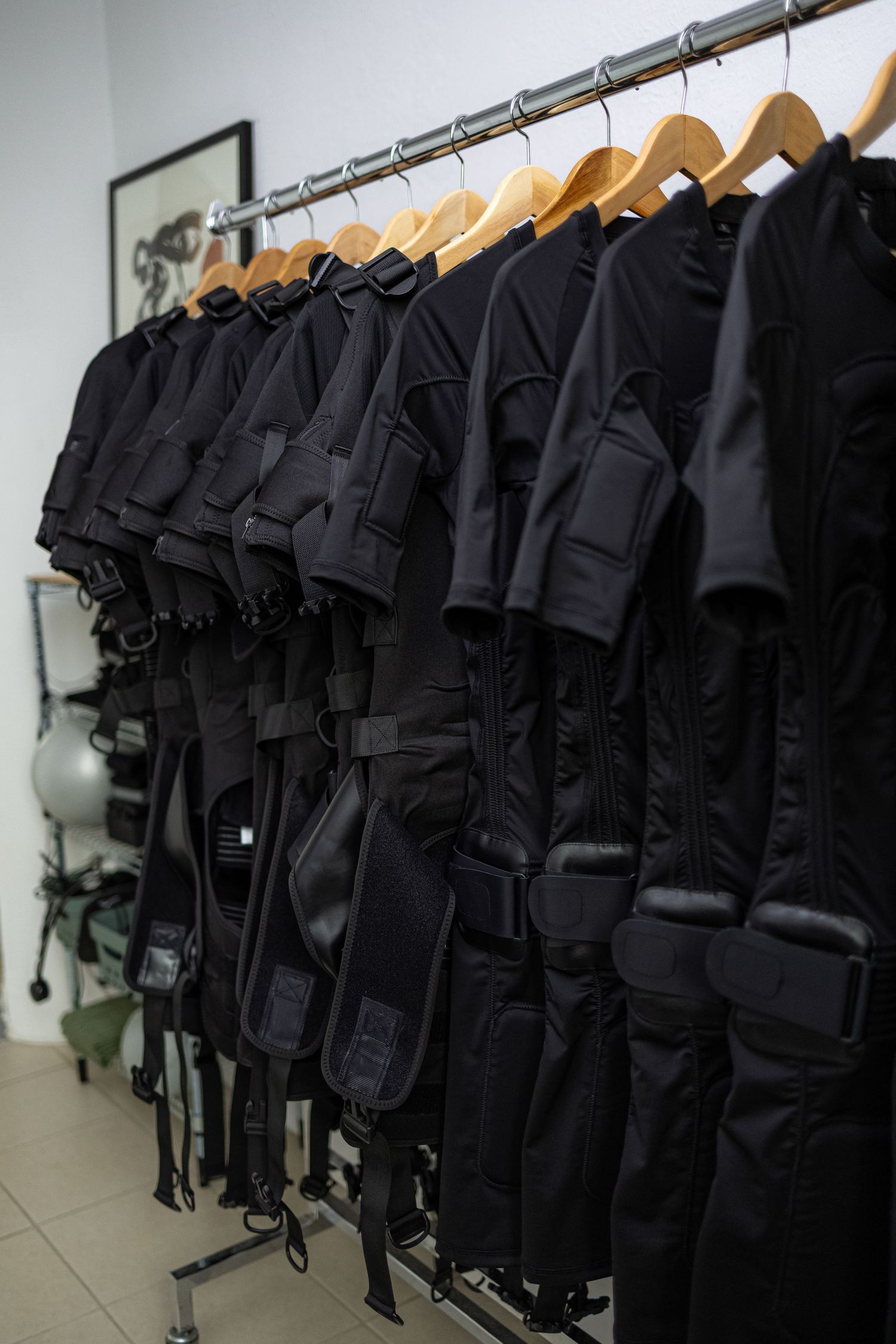 Row of black tactical vests with straps hanging on wooden hangers on a metal rack in a white room.