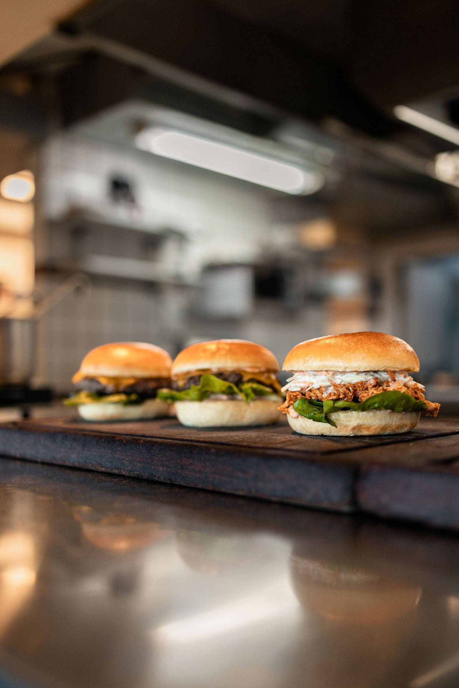 Three gourmet burgers on a wooden board in a professional kitchen, featuring fresh toppings and golden brioche buns.