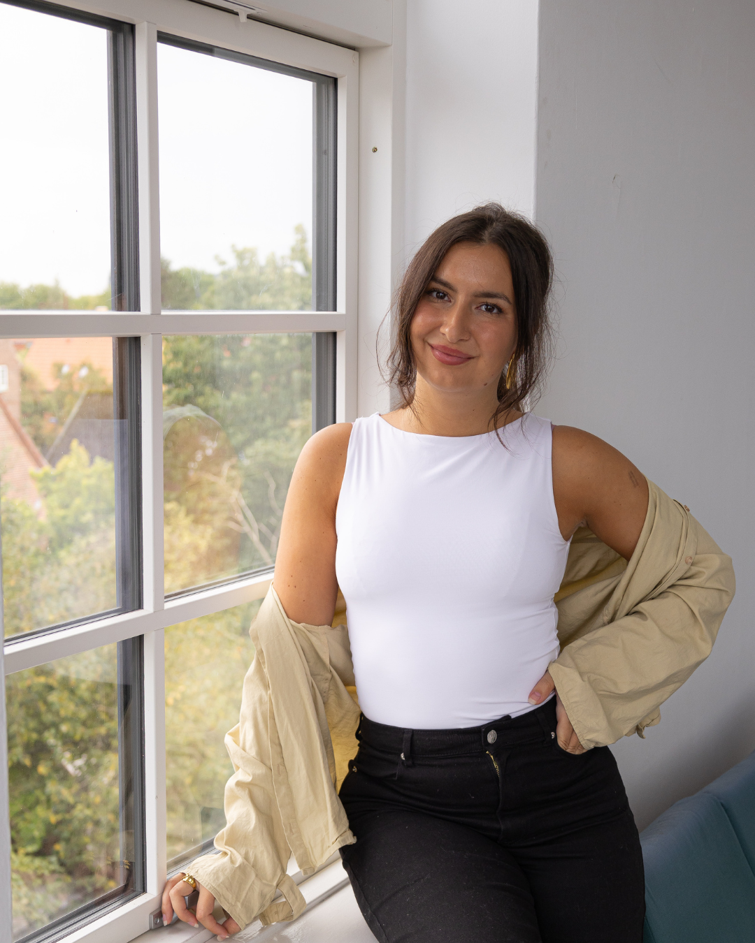 Lifestyle fashion photoshoot featuring casual white tank top and beige jacket against bright window backdrop.
