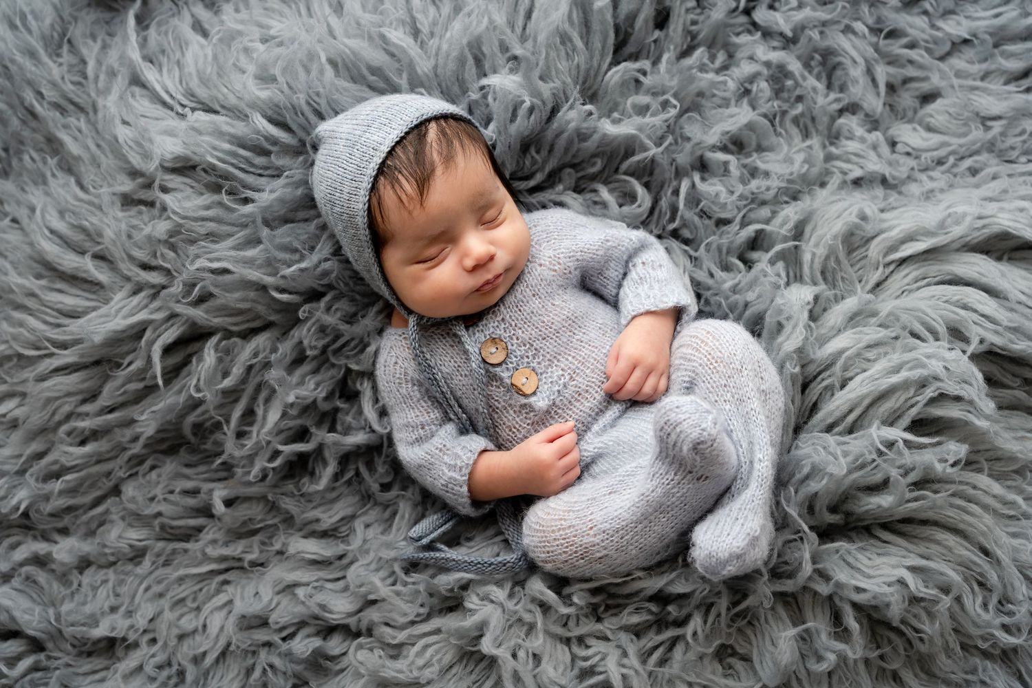 Sleeping newborn in gray outfit lying on plush gray fur blanket during professional photo session.