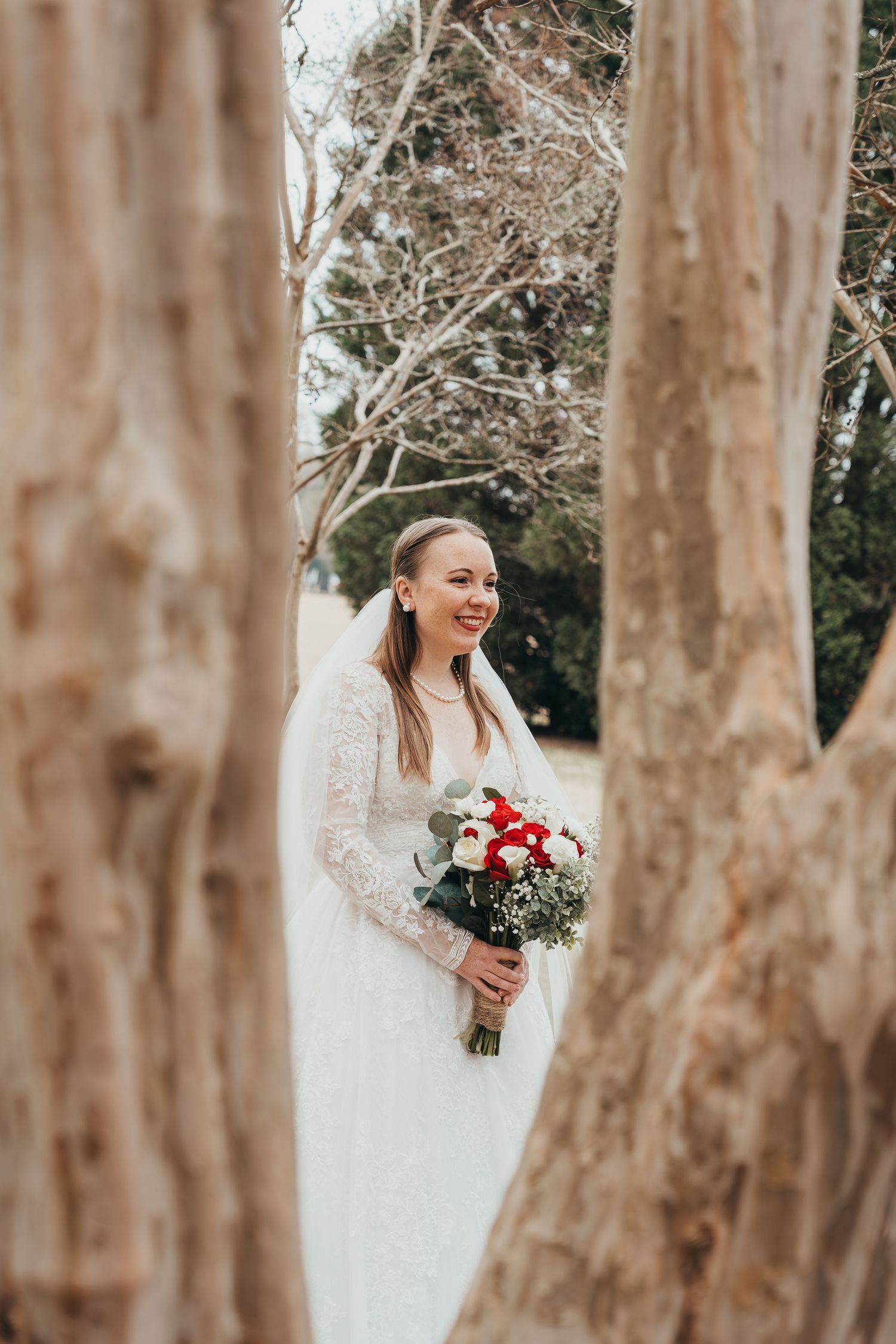 Candid bridal portrait through tree trunks showing lace dress and red bouquet.