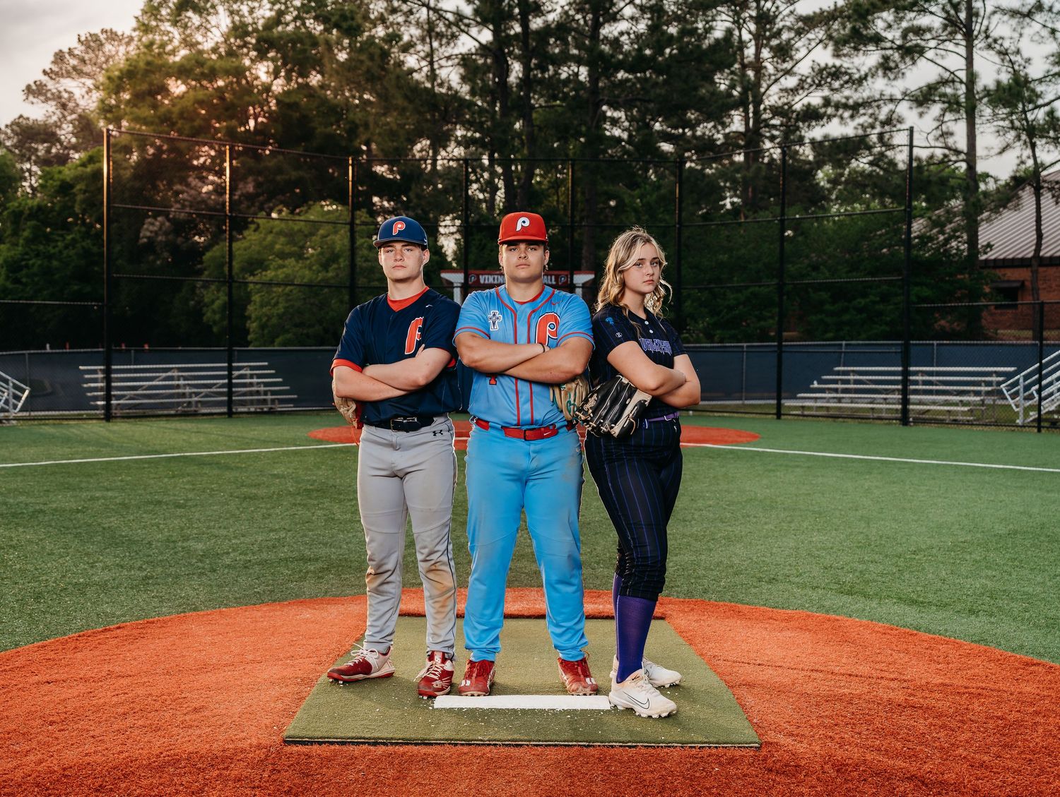 Three people pose confidently on a baseball pitcher's mound at sunset.