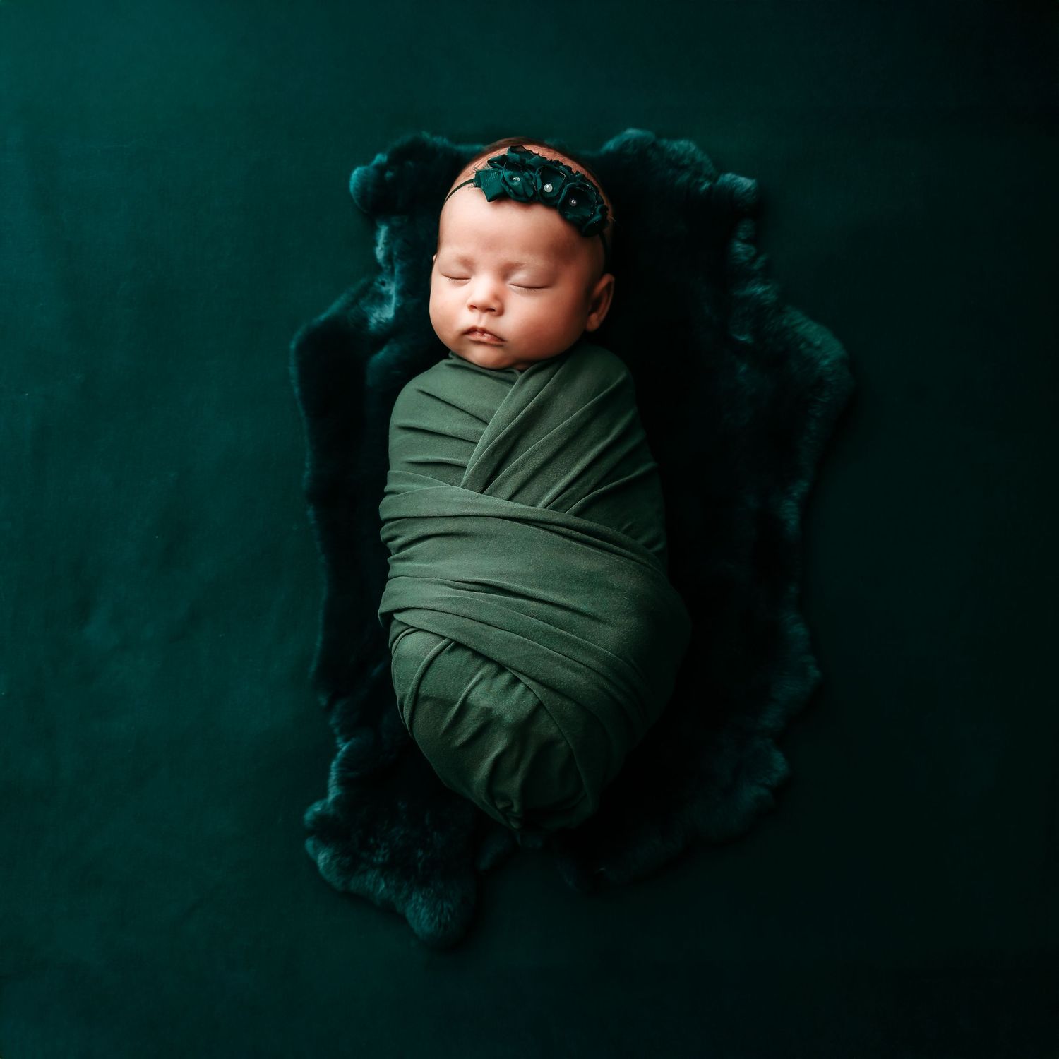 Sleeping newborn wrapped in green swaddle on dark velvet background.
