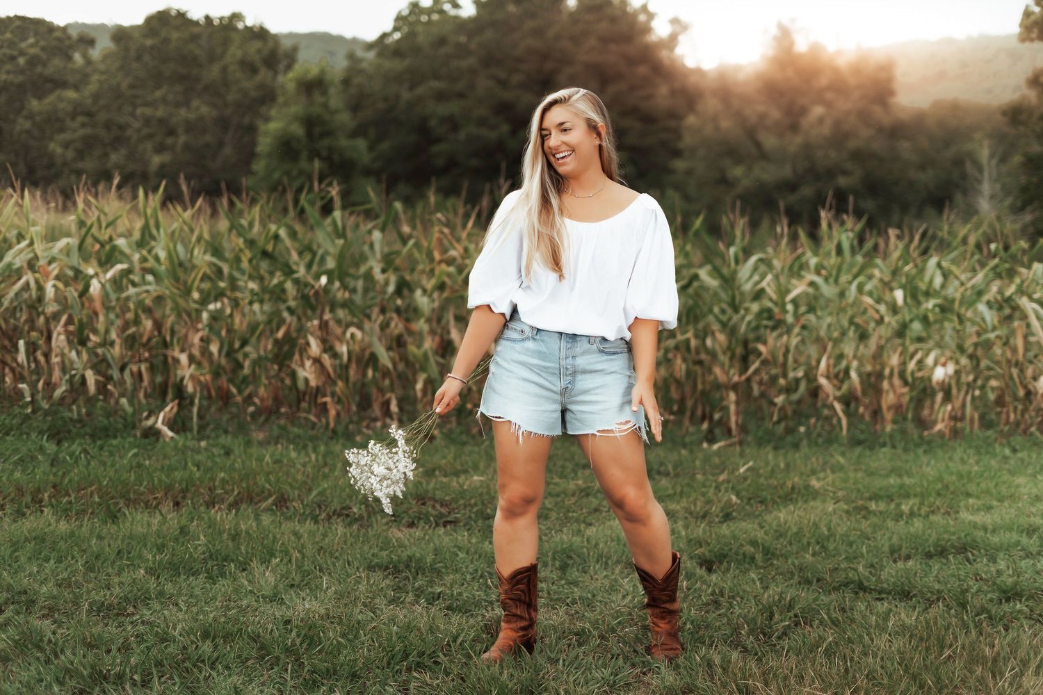 A person in a white top and denim shorts poses in a field at sunset wearing brown cowboy boots.