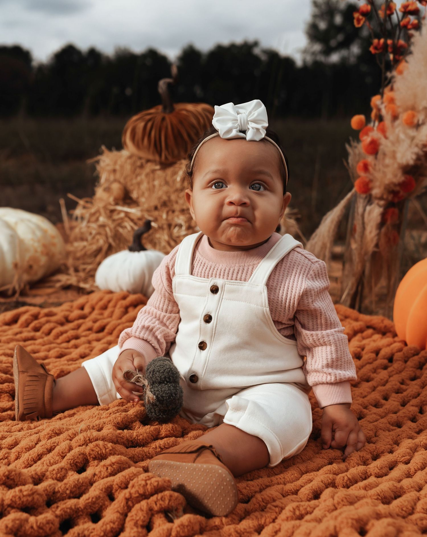 Baby in white overalls and bow sitting on orange knit blanket among fall decorations.