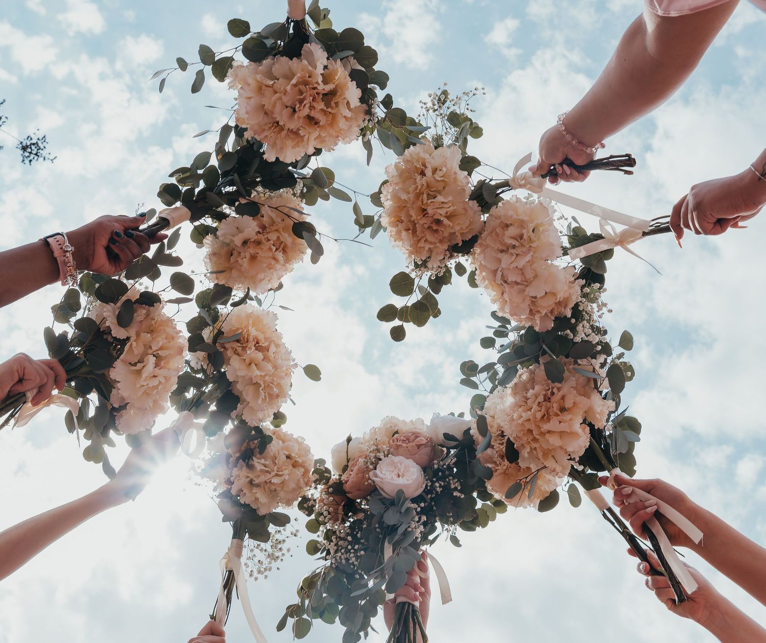 Circle of hands holding pink floral wreath against blue sky creating artistic wedding photo composition.