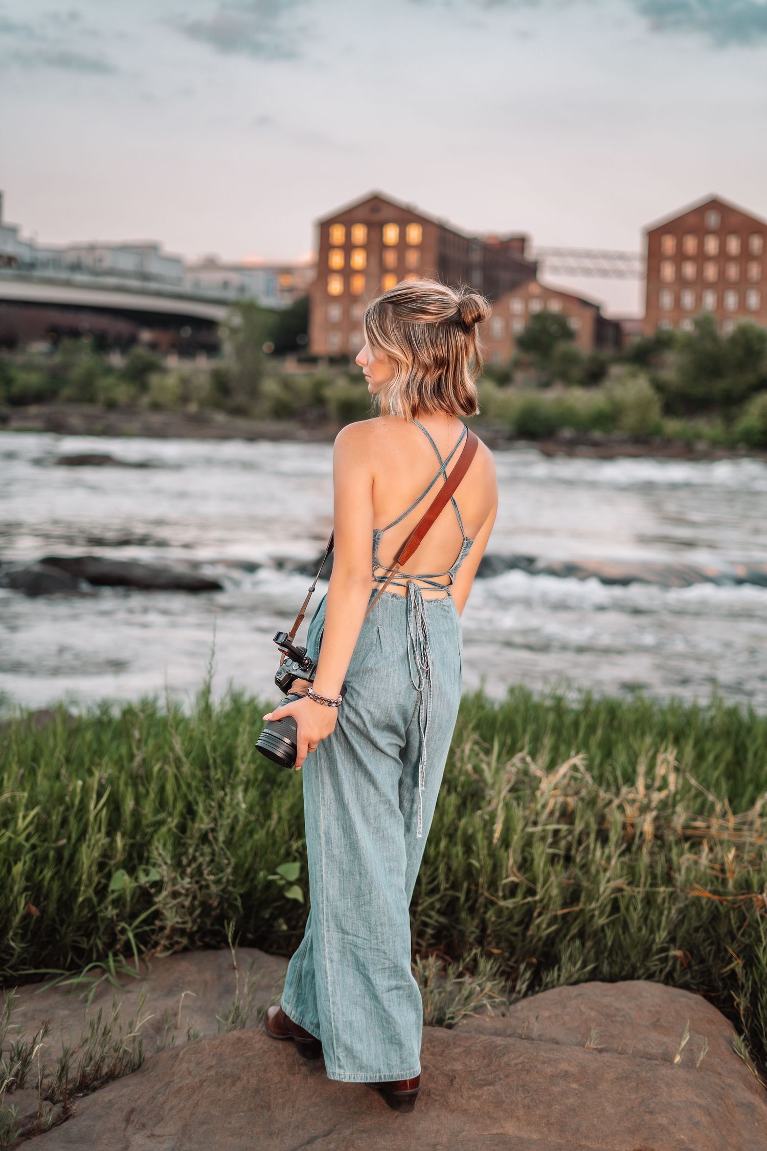 Woman in denim jumpsuit holding camera stands by river with industrial buildings in background.