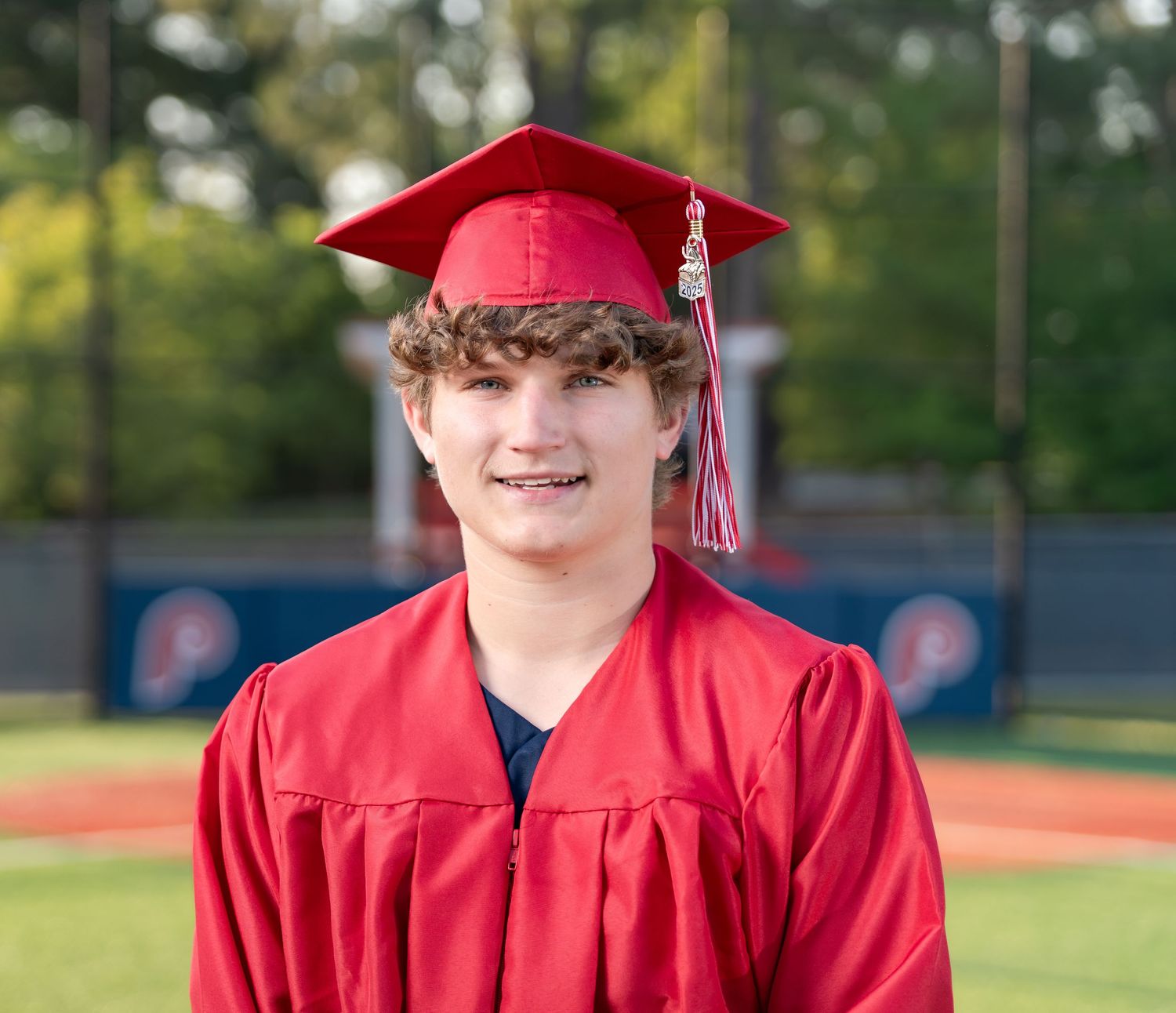 Graduate in red cap and gown smiles on baseball field during outdoor graduation photo session.