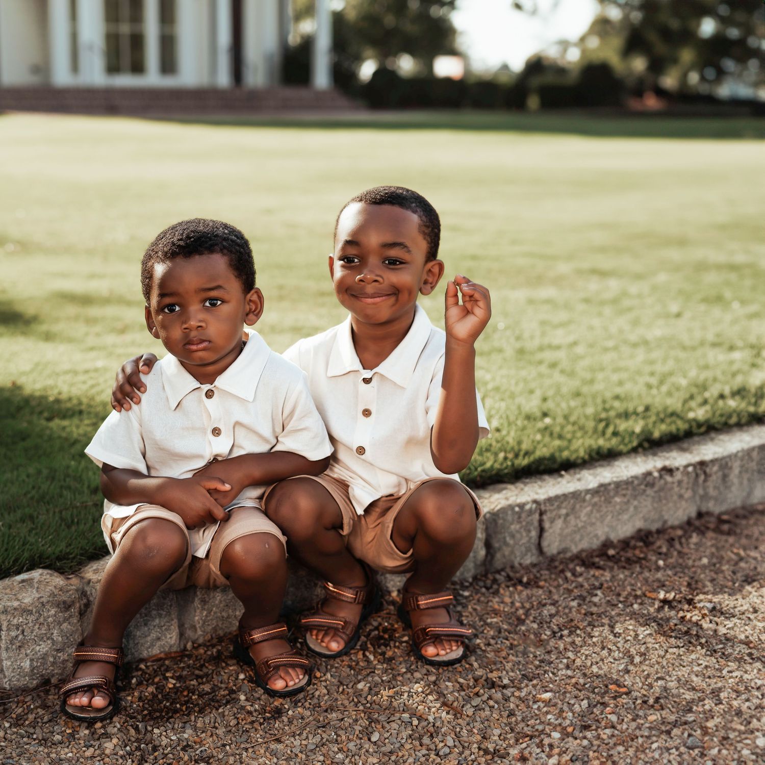 Two young children in white polo shirts sitting together on a curb wearing sandals.