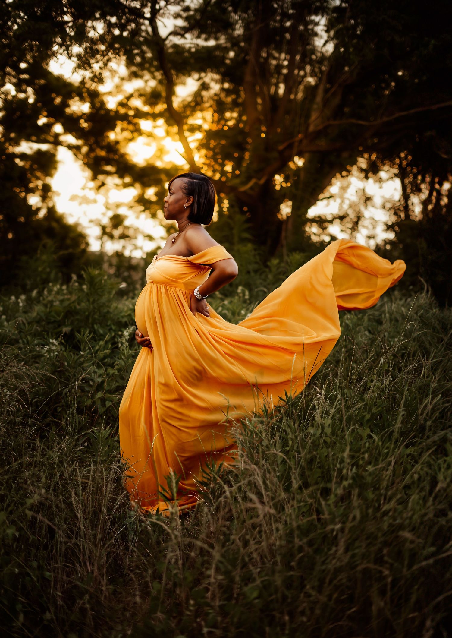 Figure in flowing yellow dress poses dramatically in golden sunset light among tall grass.