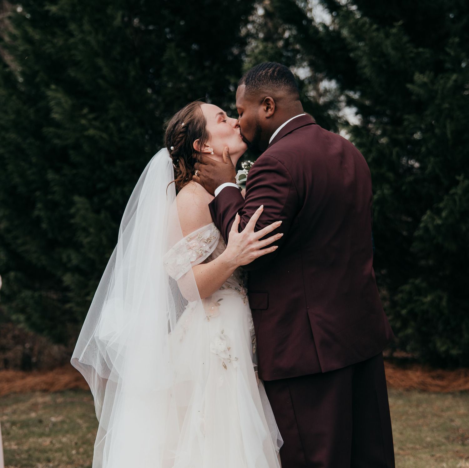 A romantic wedding moment captured outdoors with a burgundy suit and flowing white dress.