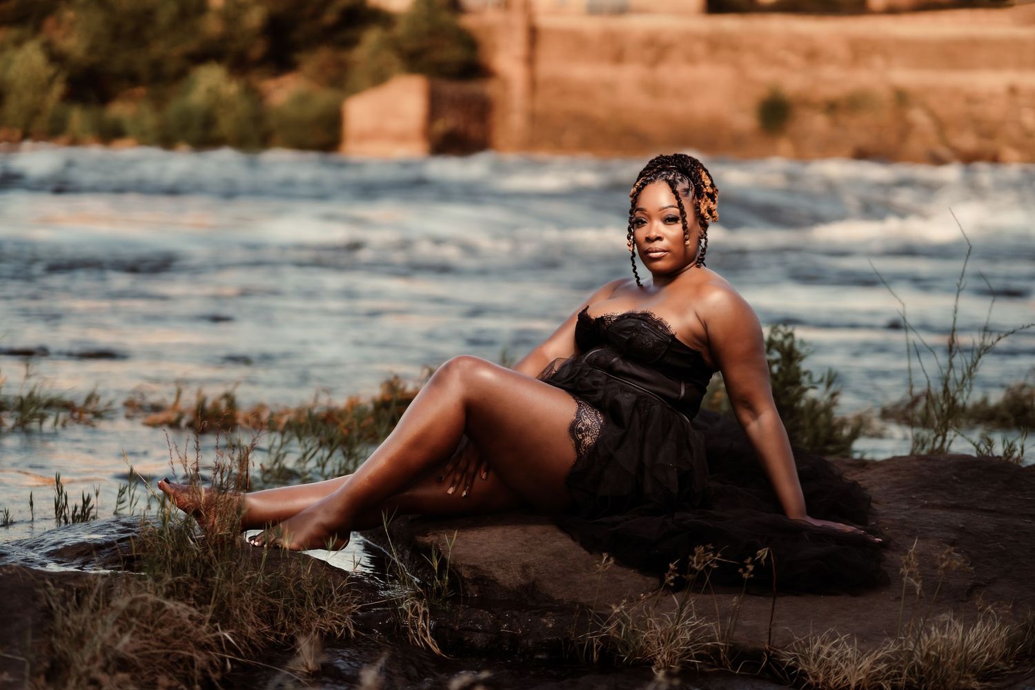 A person in a black dress poses on rocks near a flowing river during golden hour lighting.