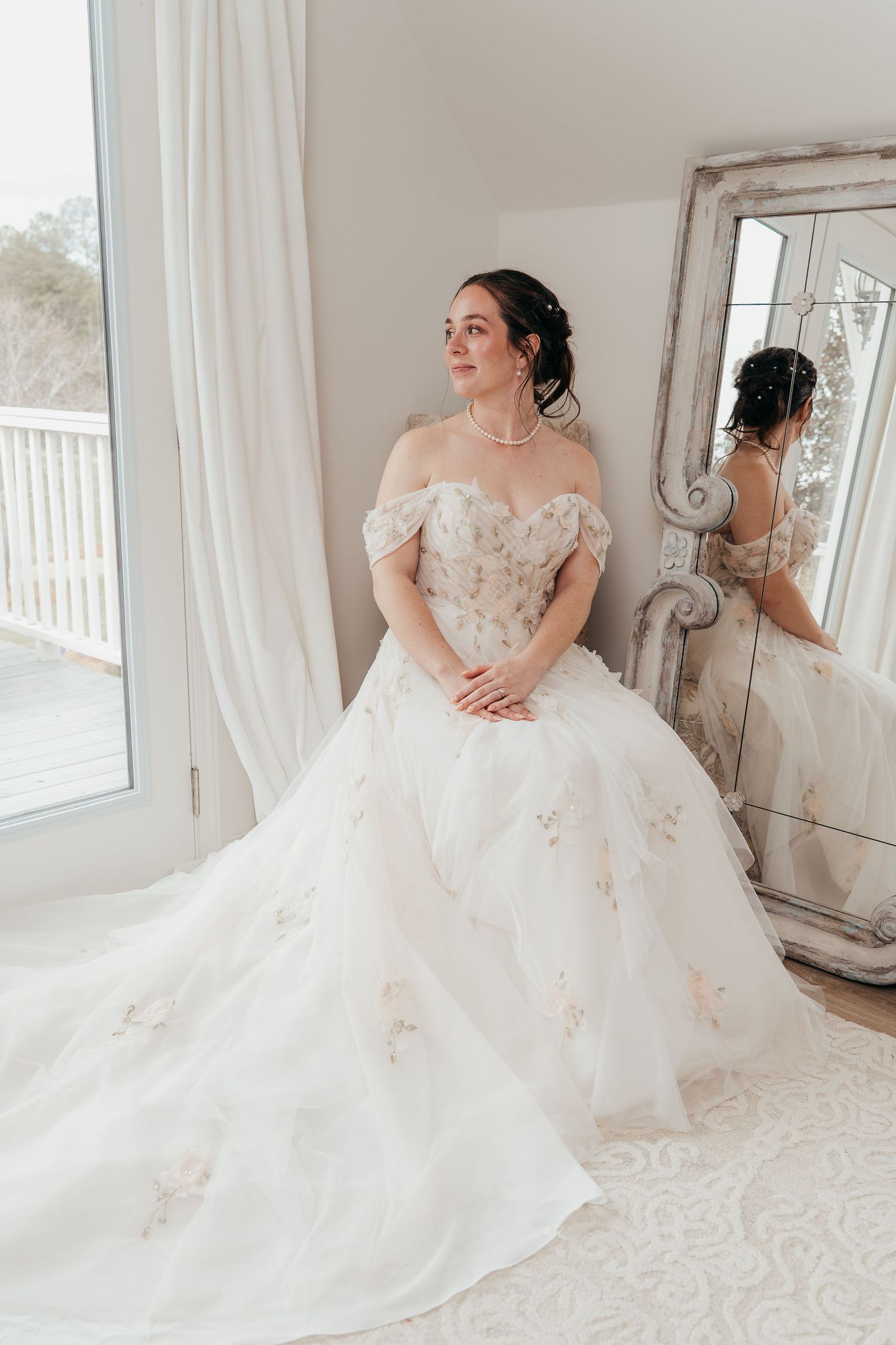 Elegant off-shoulder wedding gown with floral details photographed next to vintage mirror in natural window light.