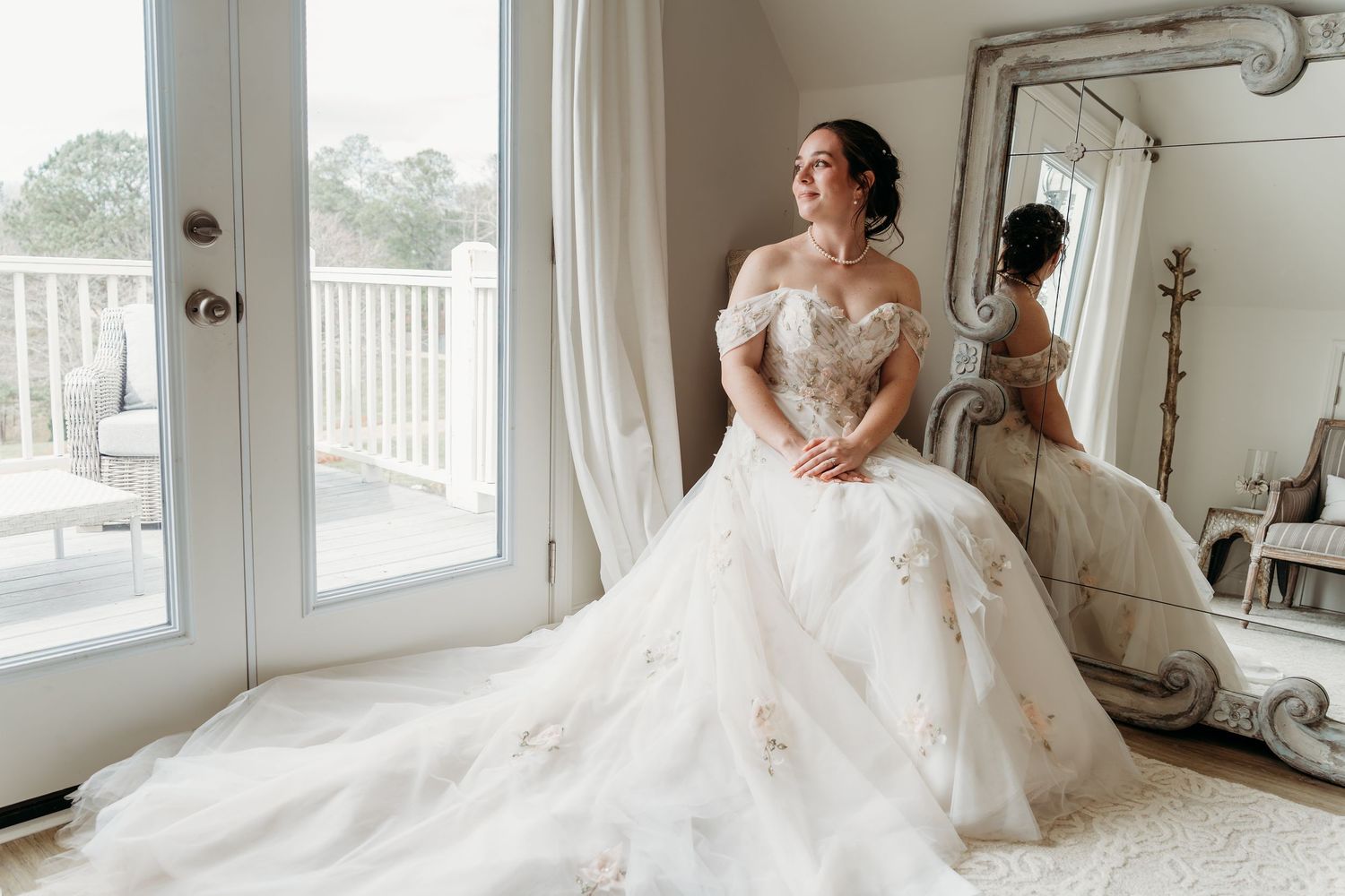 Elegant bridal gown with off-shoulder lace details and flowing train reflects in ornate mirror beside glass doors.