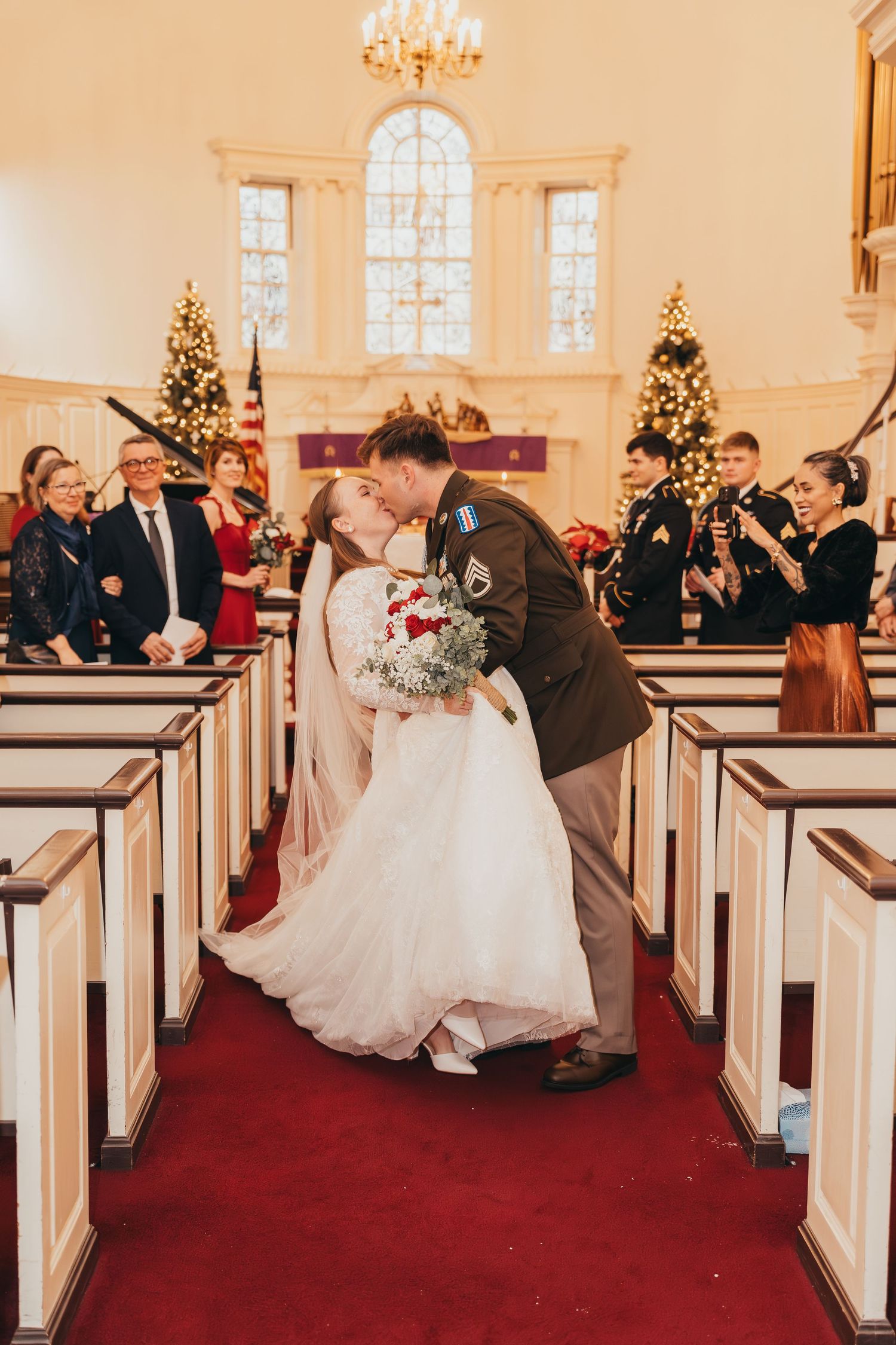 Church ceremony kiss with decorated Christmas trees and wooden pews in traditional chapel setting.