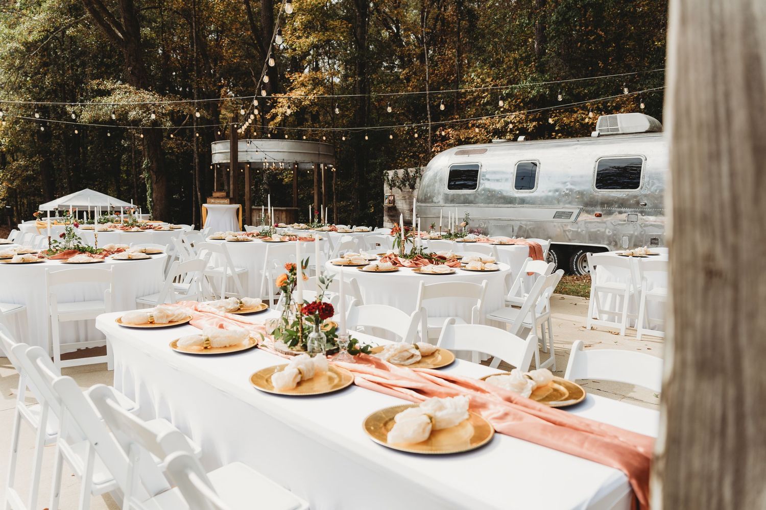 Elegant outdoor wedding reception setup with white tables and vintage airstream trailer in background.