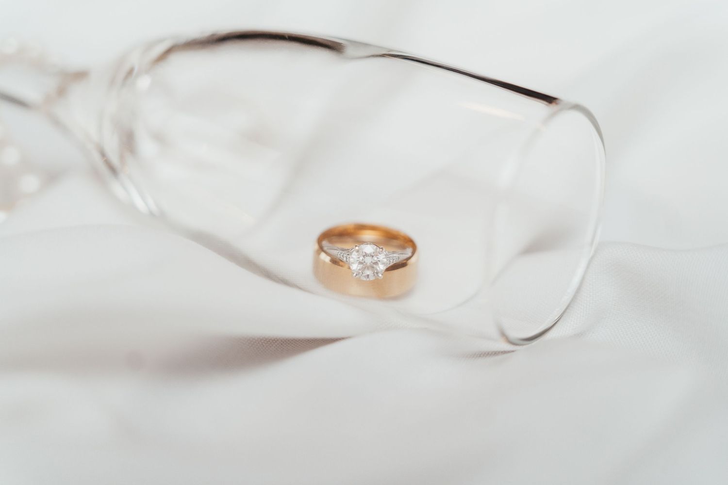 Gold wedding ring with diamond placed inside a clear wine glass on white background.