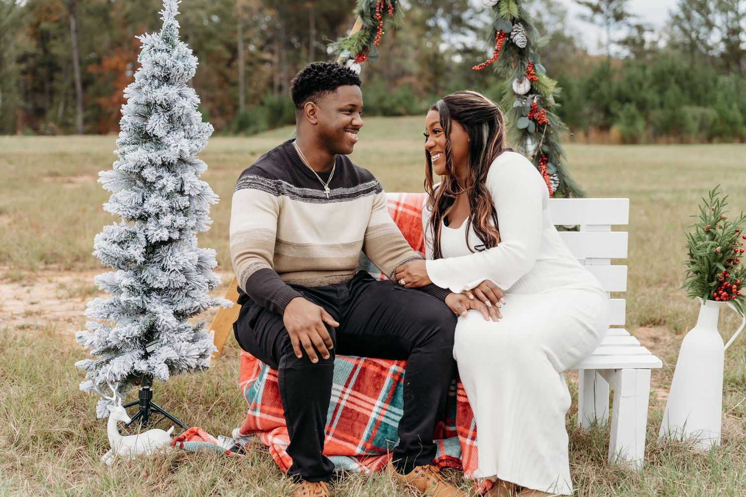 A couple sits on a white bench near a decorated Christmas tree outdoors.