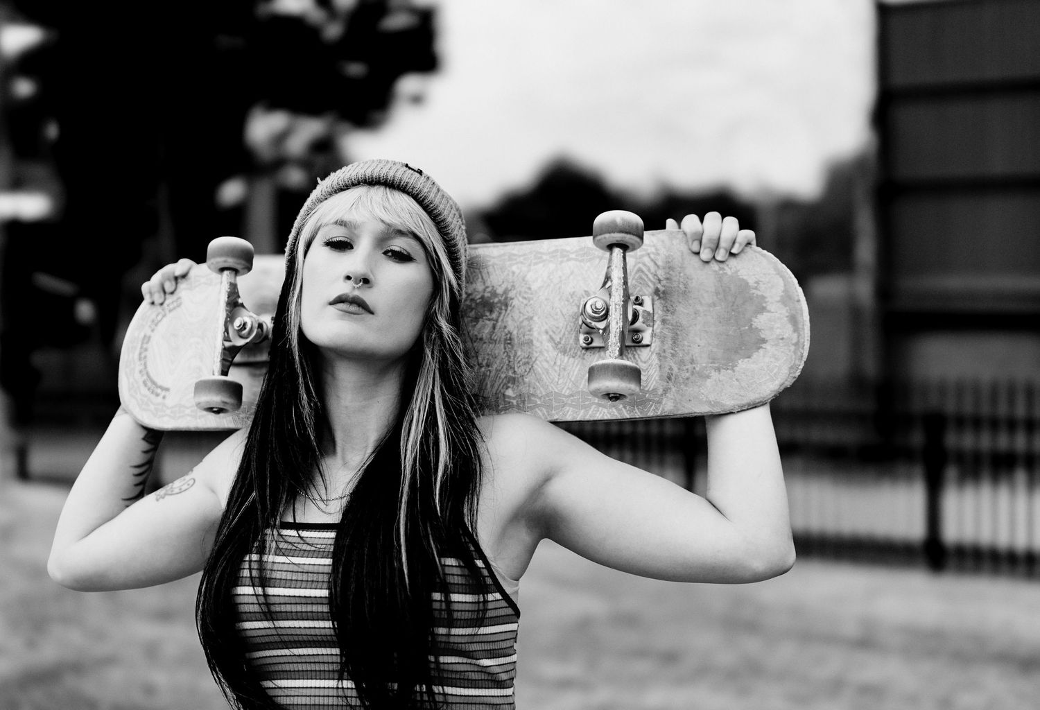 Black and white portrait of skater in striped tank top holding a skateboard behind their neck outdoors.
