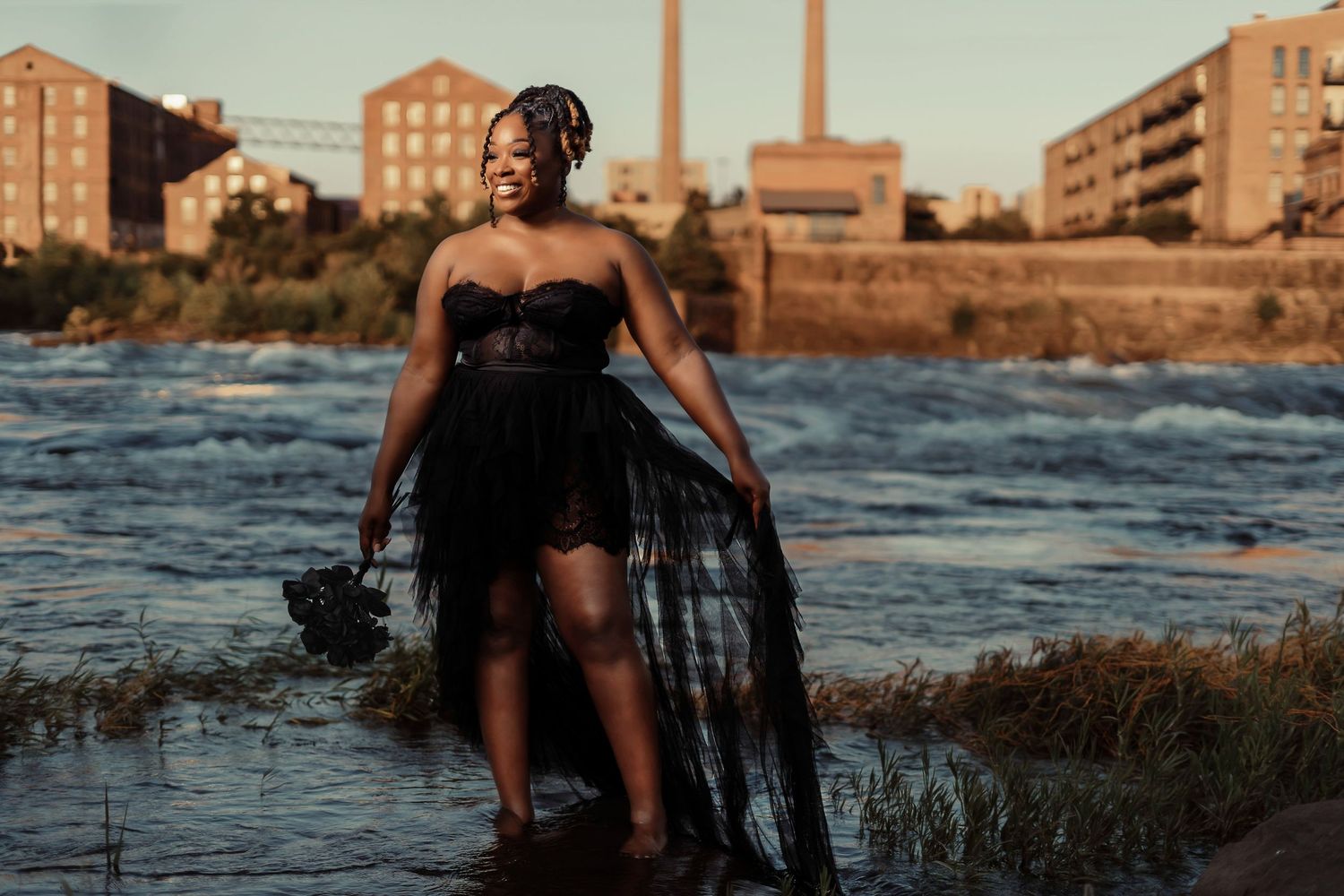 Person in flowing black dress stands in shallow water with industrial backdrop.