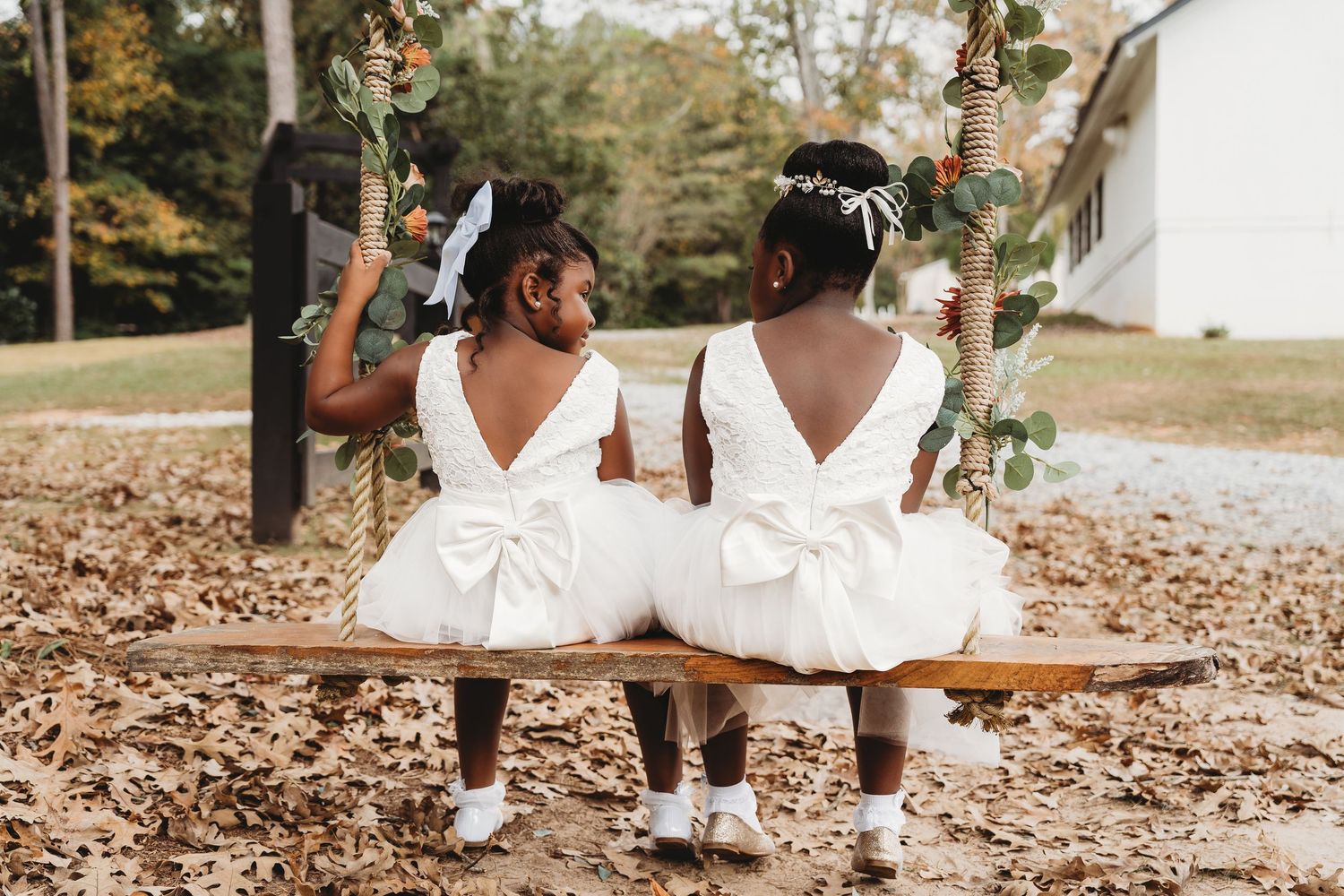 Two flower girls in white dresses with bows sitting together on a wooden swing.