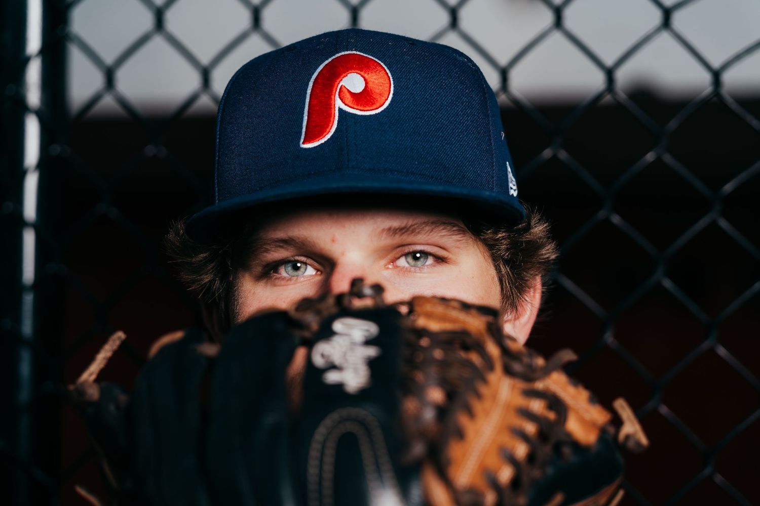 Baseball player wearing Phillies cap peeks over leather baseball glove.