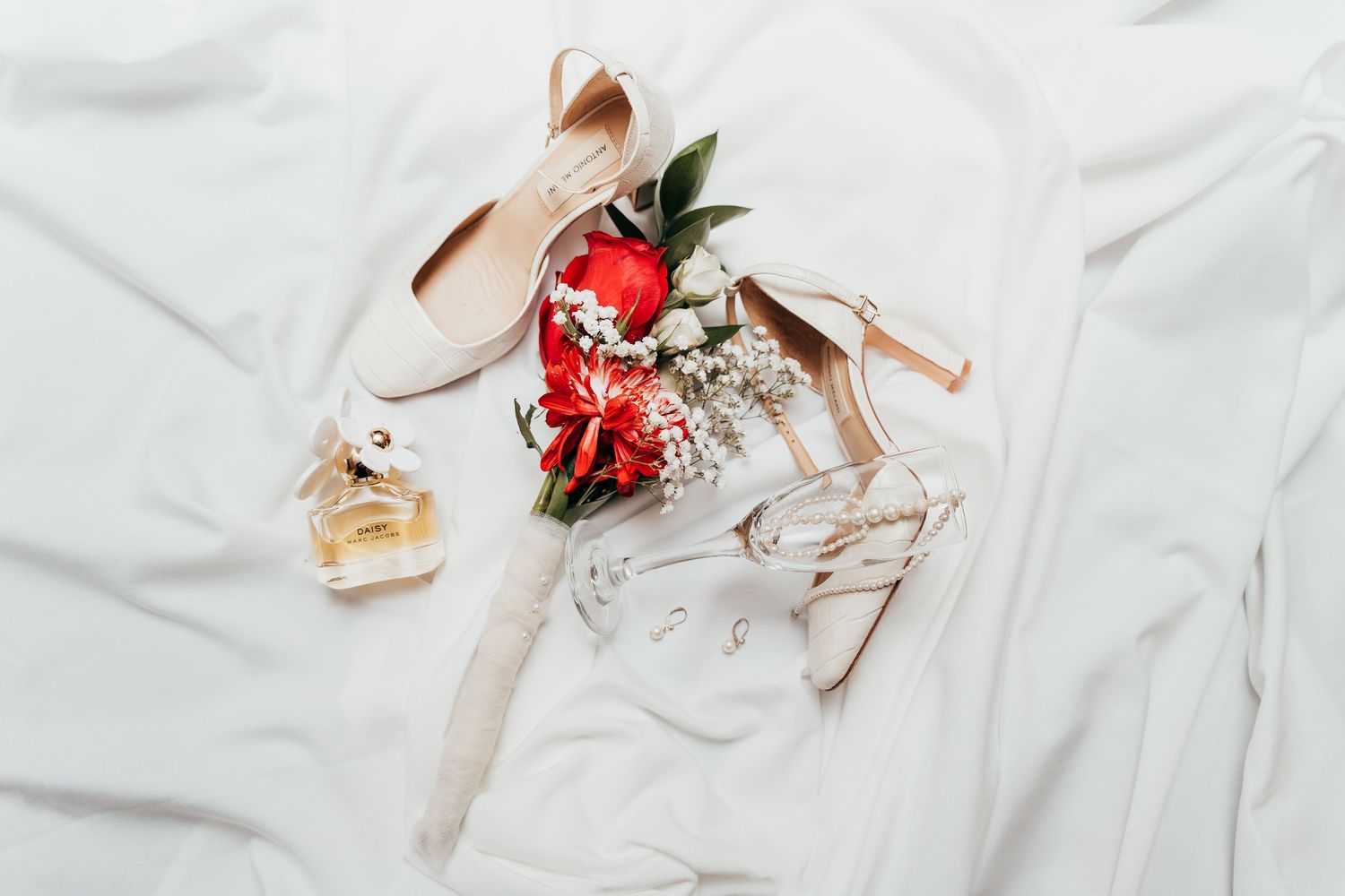 Wedding accessories including high heel shoes, perfume bottle, and red roses arranged on white fabric.