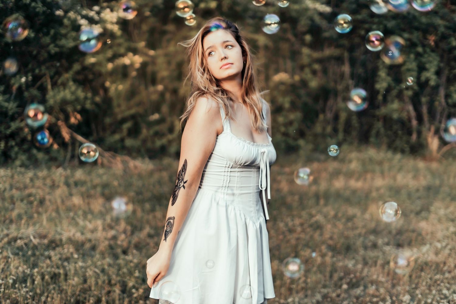 A person in a white dress stands in a meadow surrounded by floating soap bubbles on a sunny afternoon.