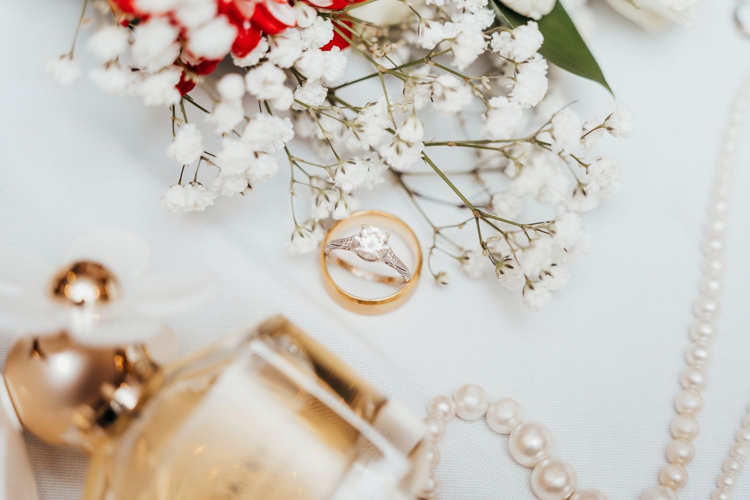 Wedding rings placed on white surface with delicate baby's breath flowers, red blooms, pearls and gold decorative elements.