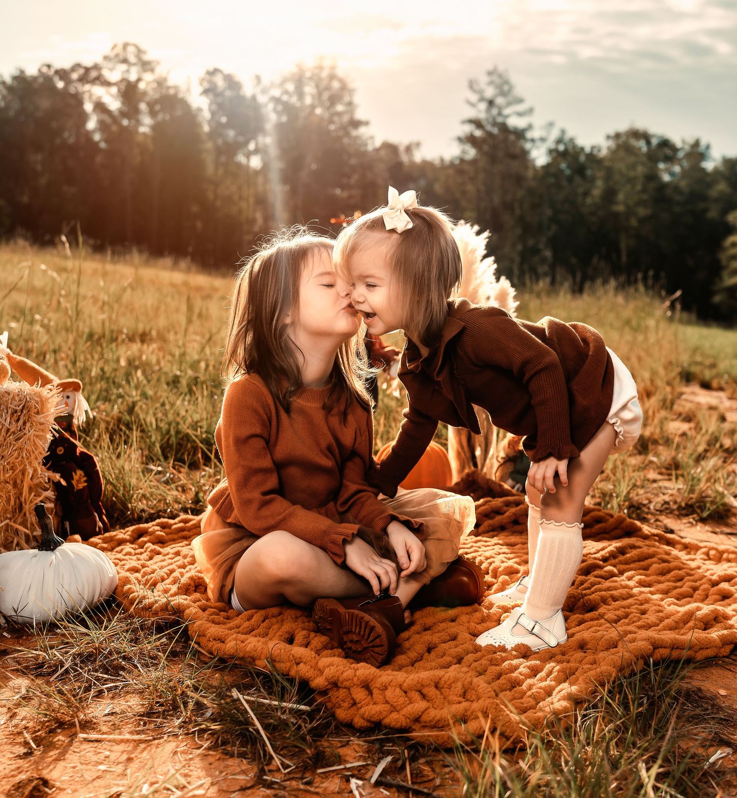 Two siblings sharing a tender moment on a rustic orange blanket in a sunlit autumn field.