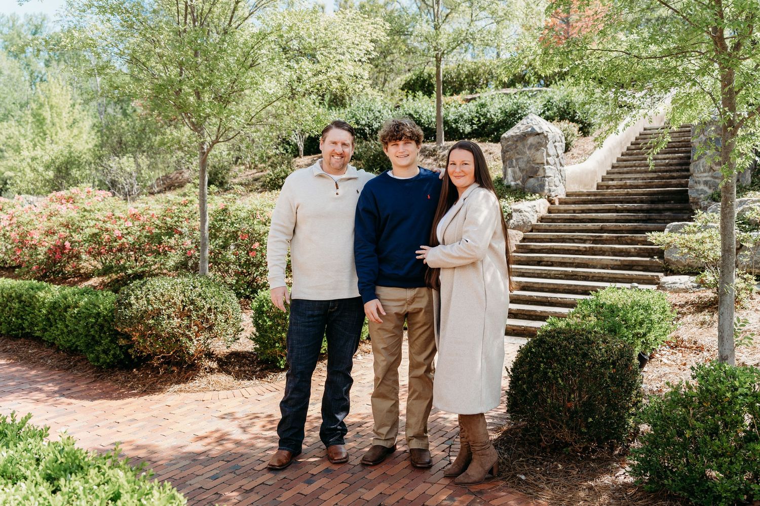 Three people stand together on a garden path with brick stairs and landscaping in the background.