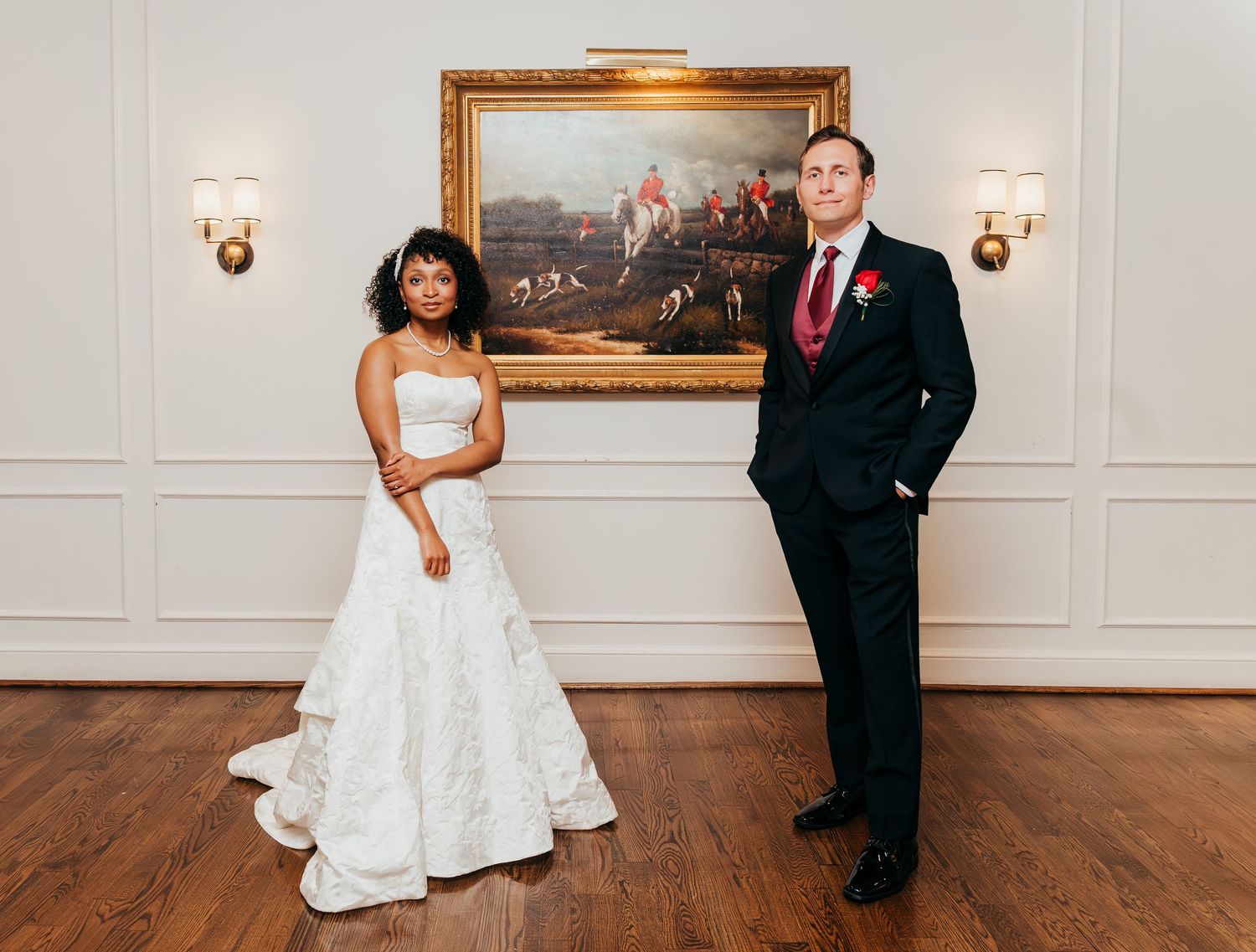 A couple poses formally in wedding attire in front of a framed painting on an elegant white wall with sconces.