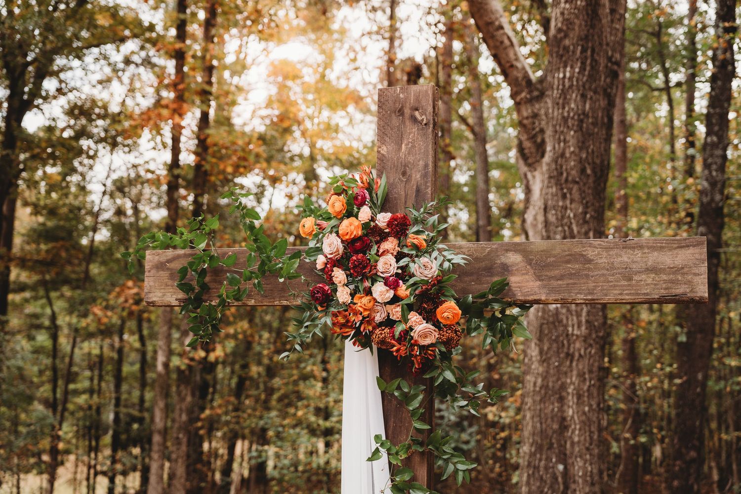 Rustic wooden cross adorned with vibrant autumn flowers and greenery in a forest setting at sunset.