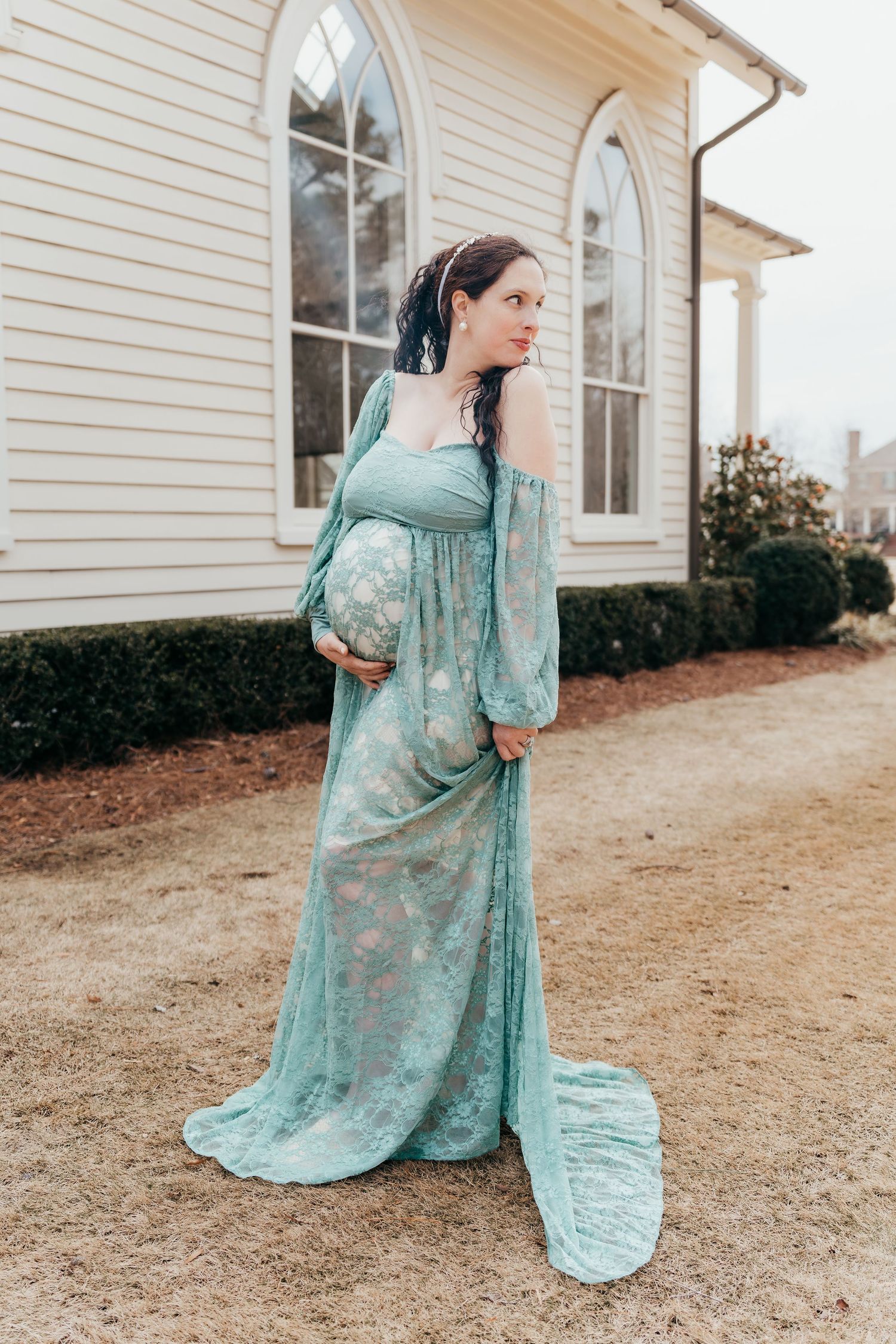 A pregnant woman in an elegant teal lace maternity gown poses in front of a white church building with arched windows.
