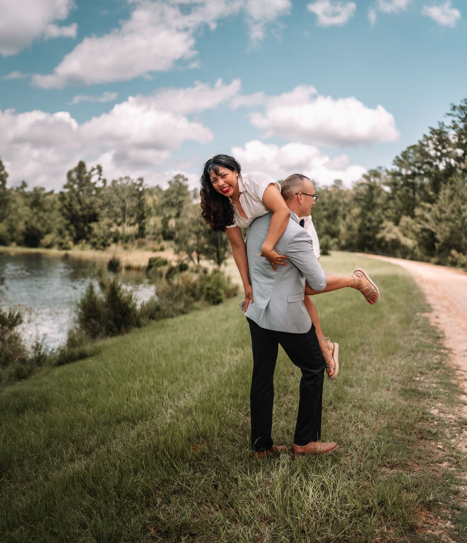 Playful couple enjoying a piggyback ride along lakeside path on sunny day.